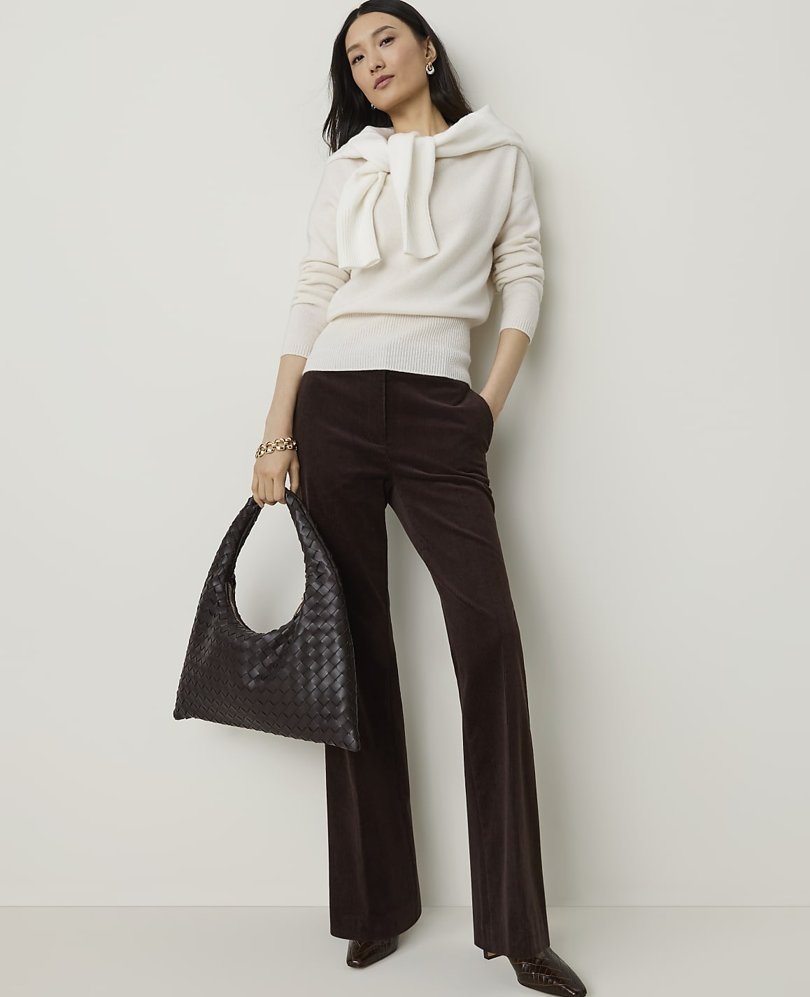 A woman in a cream sweater and brown corduroy pants stands with a dark woven bag against a plain white background.