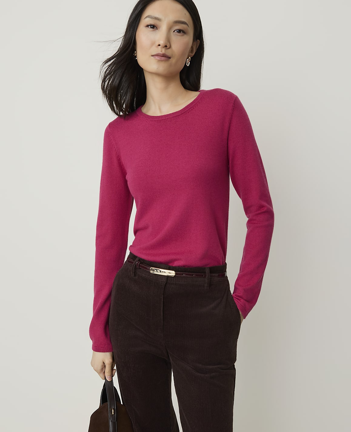 A woman in a pink sweater and brown corduroy pants stands against a plain white background, holding a dark bag.