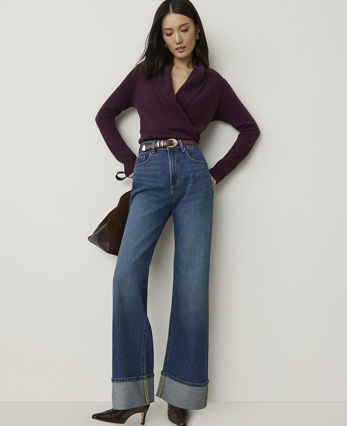 A woman in a plum-colored wrap sweater and wide-leg jeans stands against a plain white background.