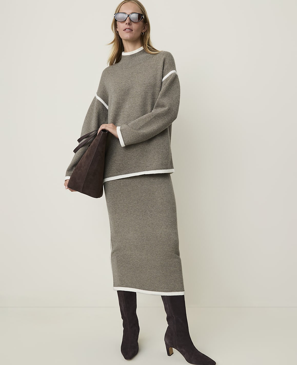 A woman in a taupe knit sweater and skirt with white trim, holding a brown suede tote bag, stands against a plain, light-colored background.