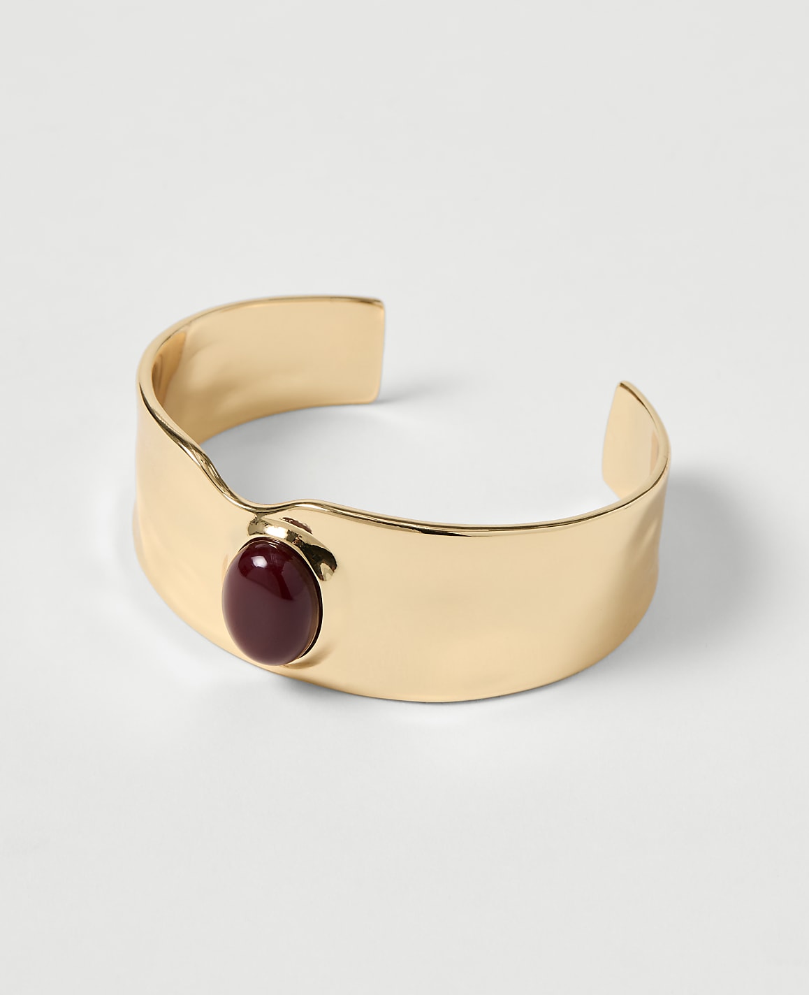 A gold cuff bracelet with a deep red stone sits on a plain white background.