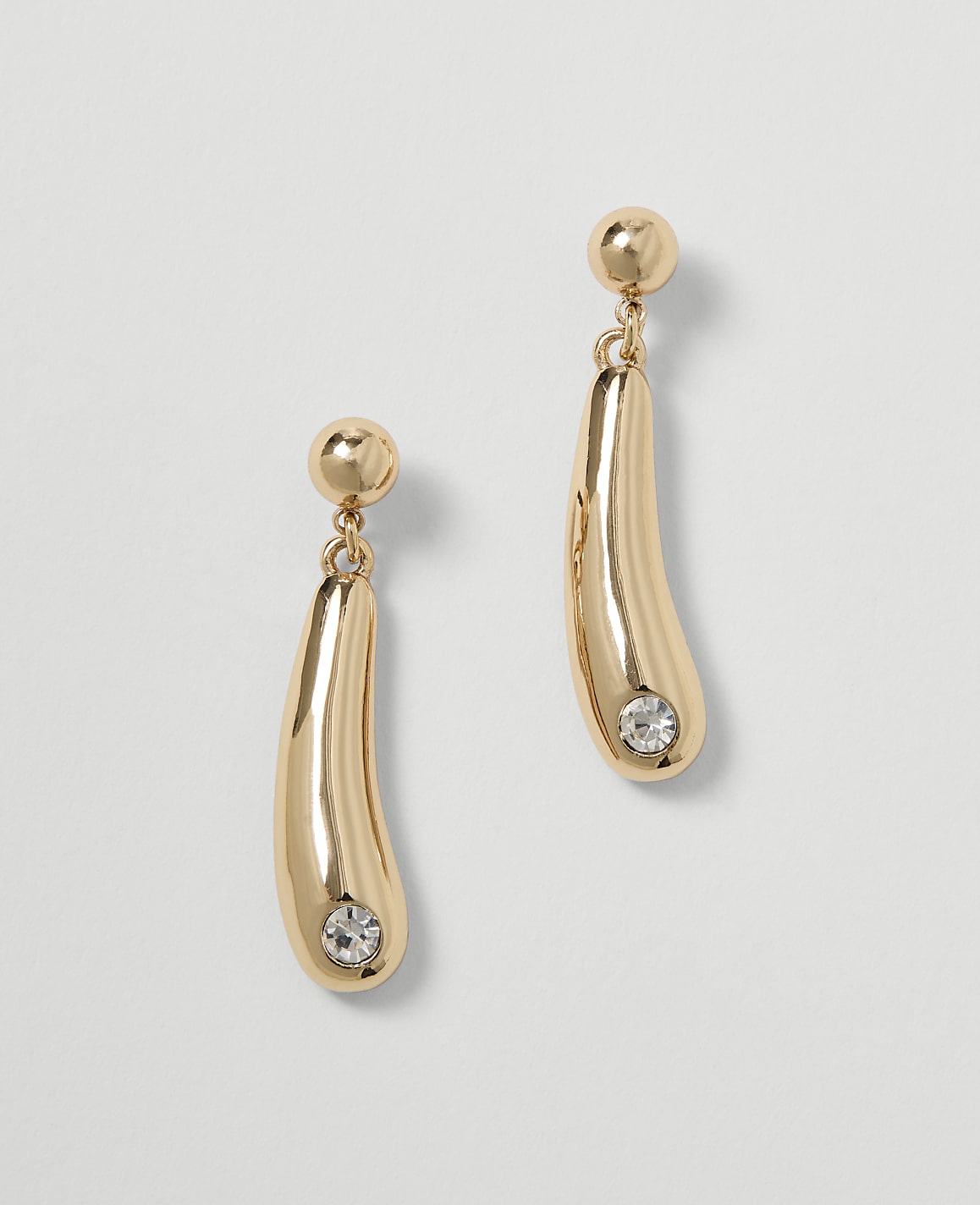 A pair of gold drop earrings with a clear gem at the bottom hang against a plain, light gray background.