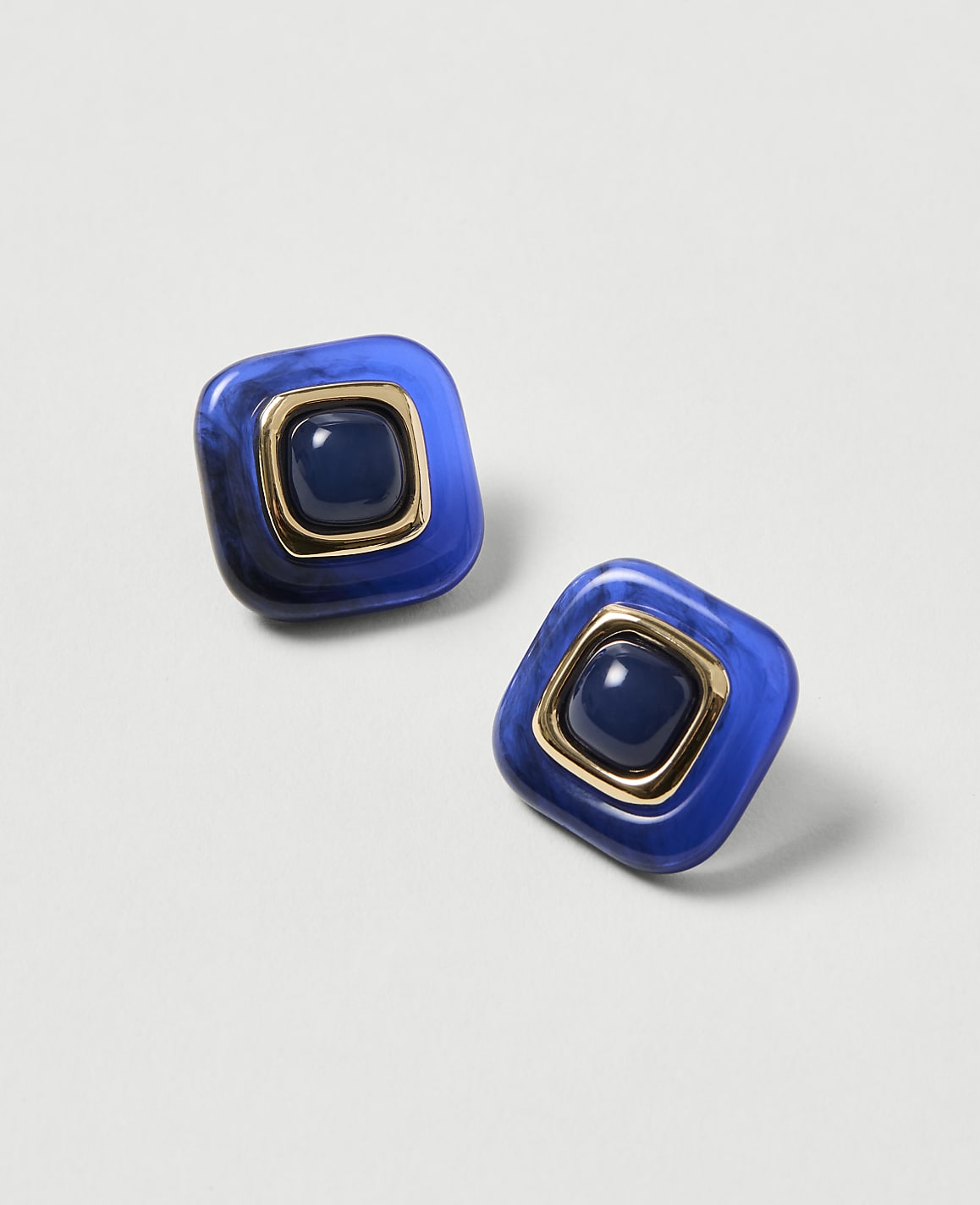 Two square blue earrings with gold trim and a dark blue center stone rest on a light gray background.