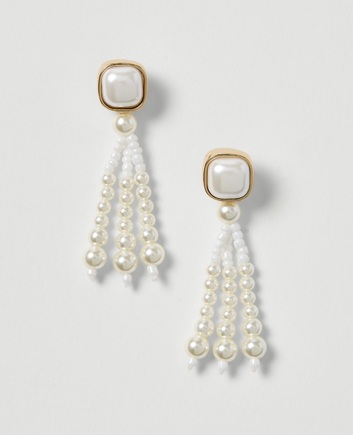 A pair of elegant pearl drop earrings with gold-toned square tops hang against a plain white background.