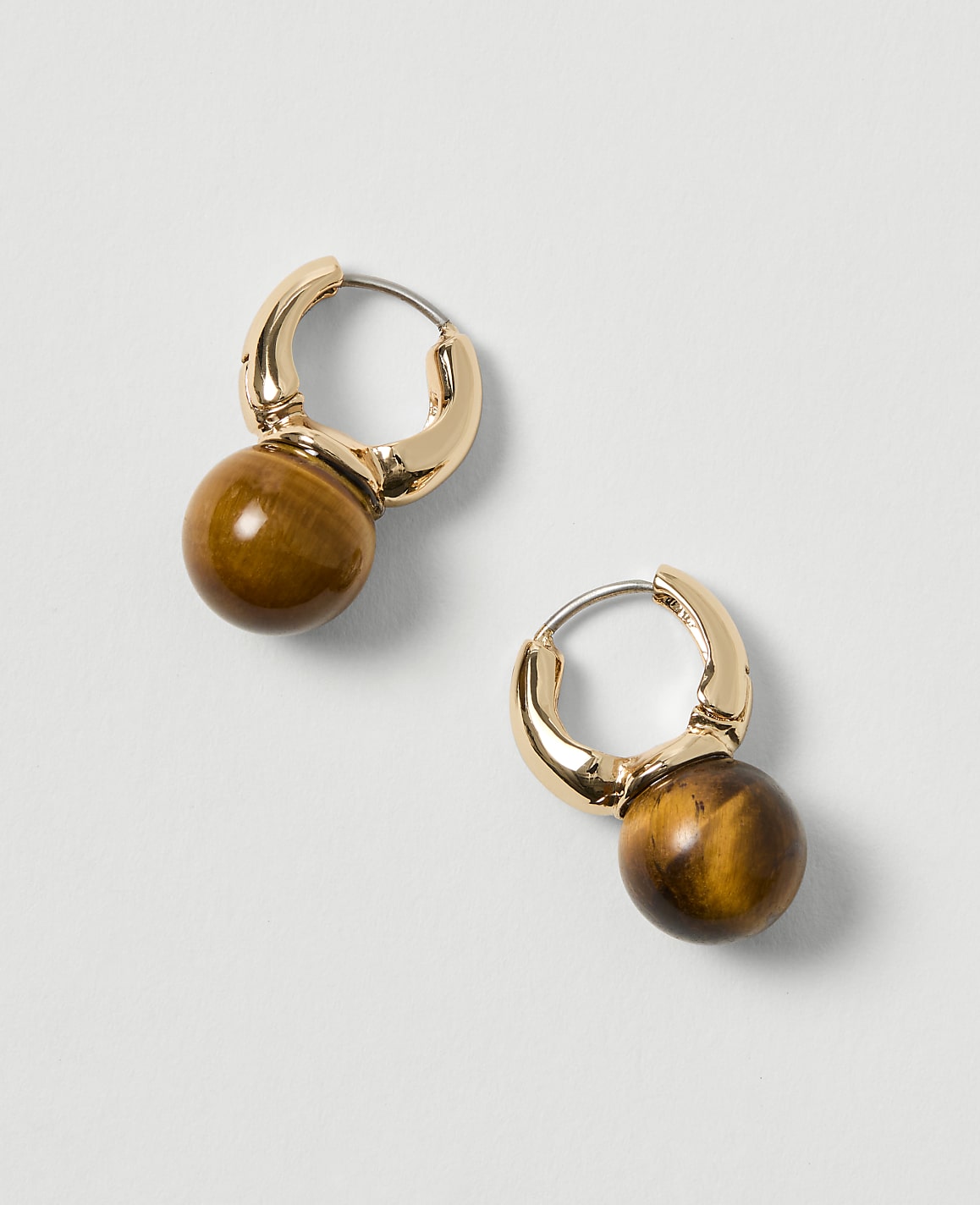 A pair of gold hoop earrings with tiger's eye beads hang against a plain, light gray background.