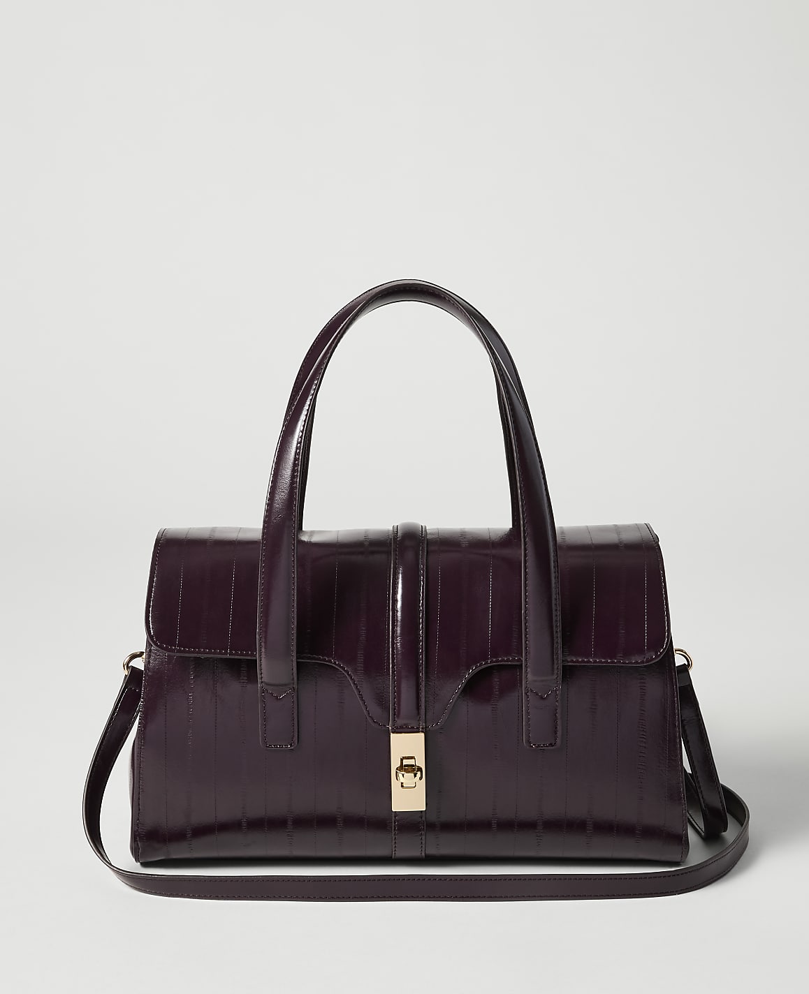 A deep purple, croc-embossed handbag with a gold clasp and top handles sits in the foreground against a plain, light gray background.