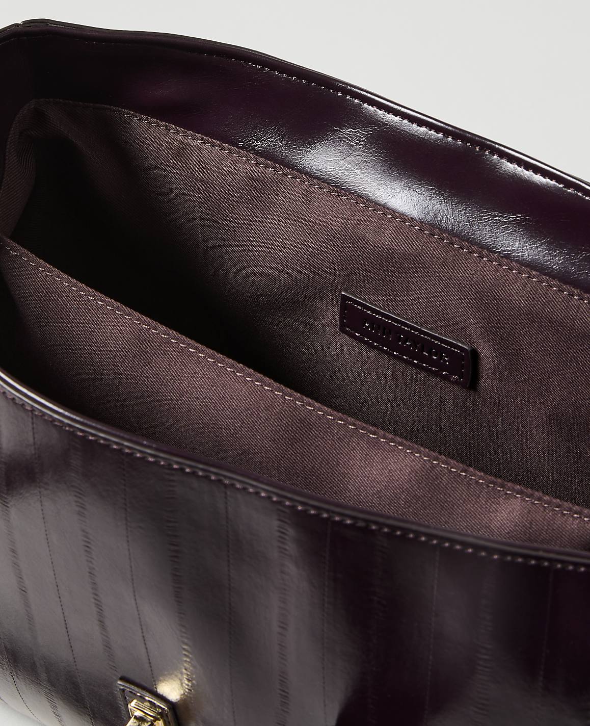 A glossy, dark purple handbag with vertical stitching is open in the foreground, revealing its brown fabric lining and a small brand tag, against a plain, light background.