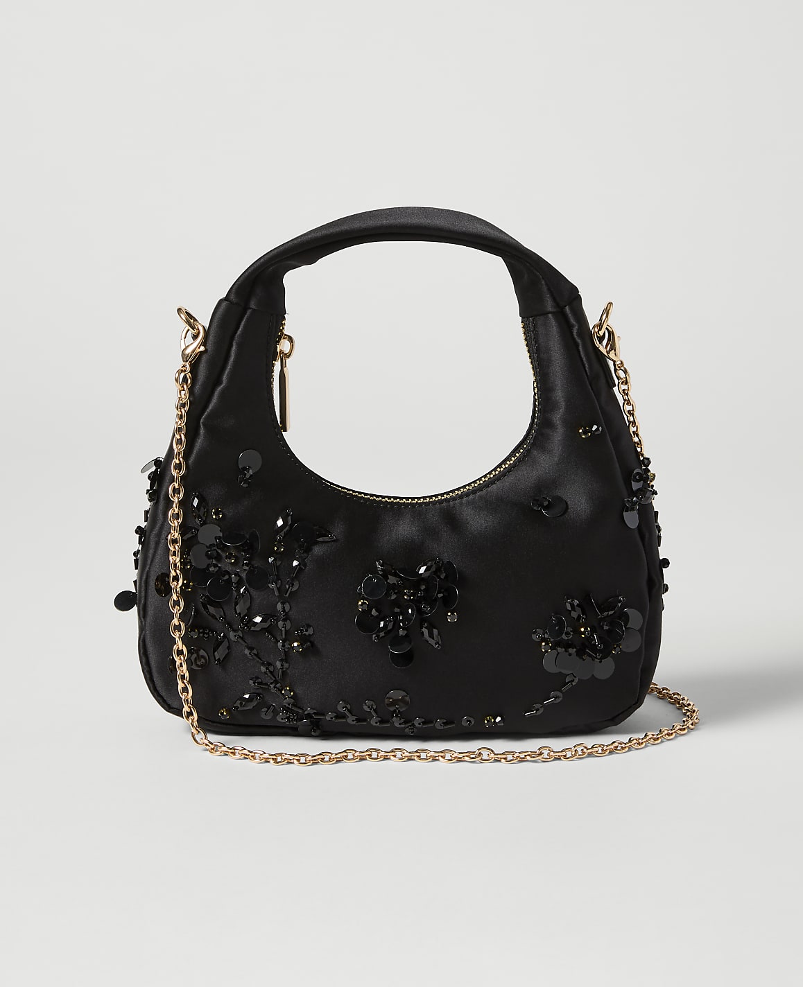A black satin handbag adorned with black sequins and beads sits on a light gray background, with a delicate gold chain strap draped across it.
