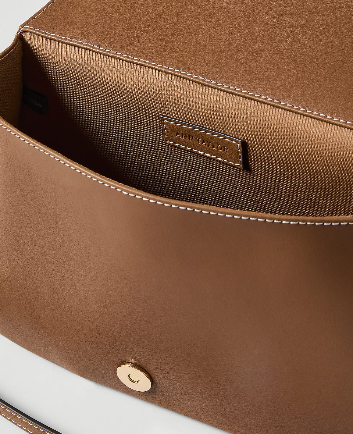 A brown leather clutch with a gold clasp is open, revealing its satin interior and a brand tag, against a plain white background.