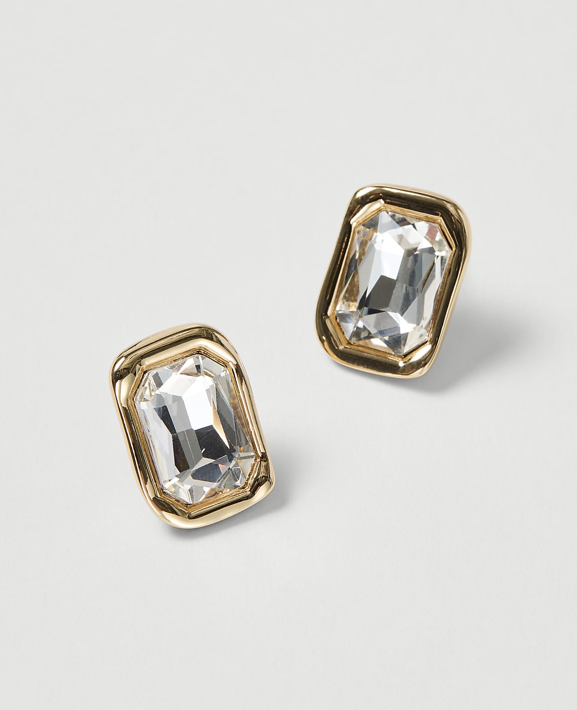 Two rectangular, gold-framed earrings with clear, faceted gemstones are displayed against a plain, light gray background.