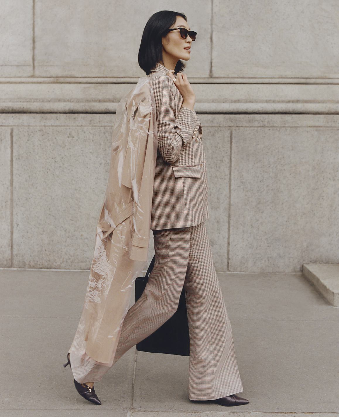 A woman in a tan plaid suit and sunglasses walks with a sheer trench coat draped over her shoulder in front of a stone building.