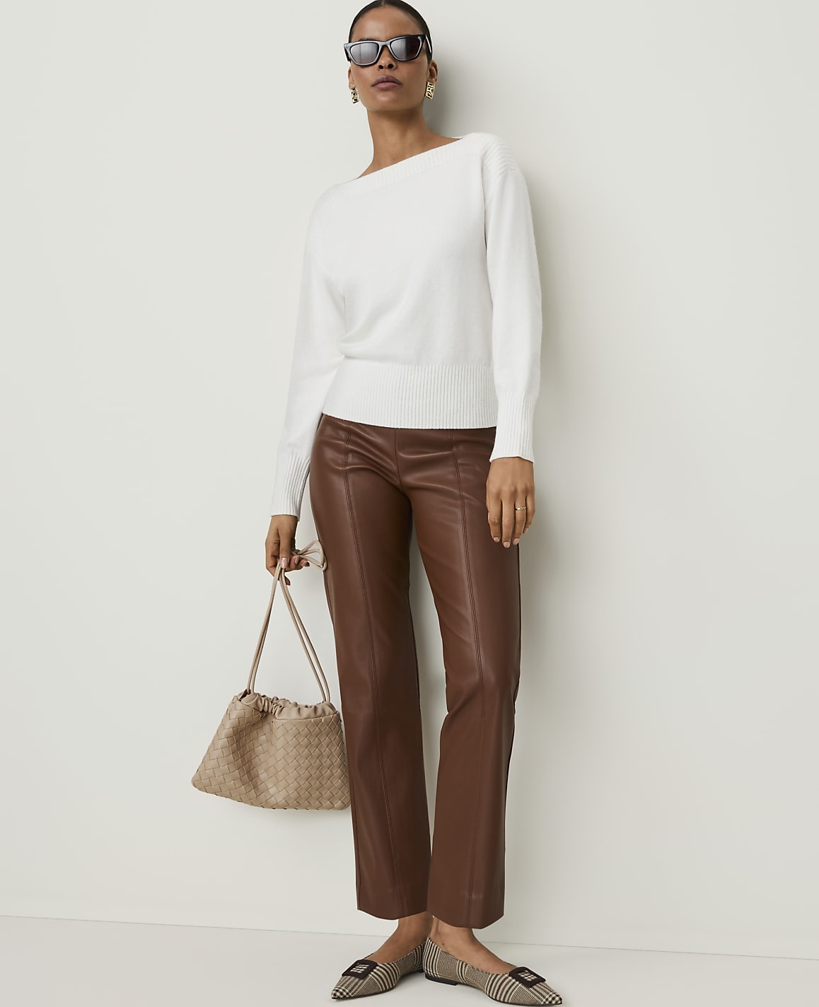 A woman in a white sweater and brown leather pants stands against a plain white background, holding a beige handbag and wearing checkered flats.