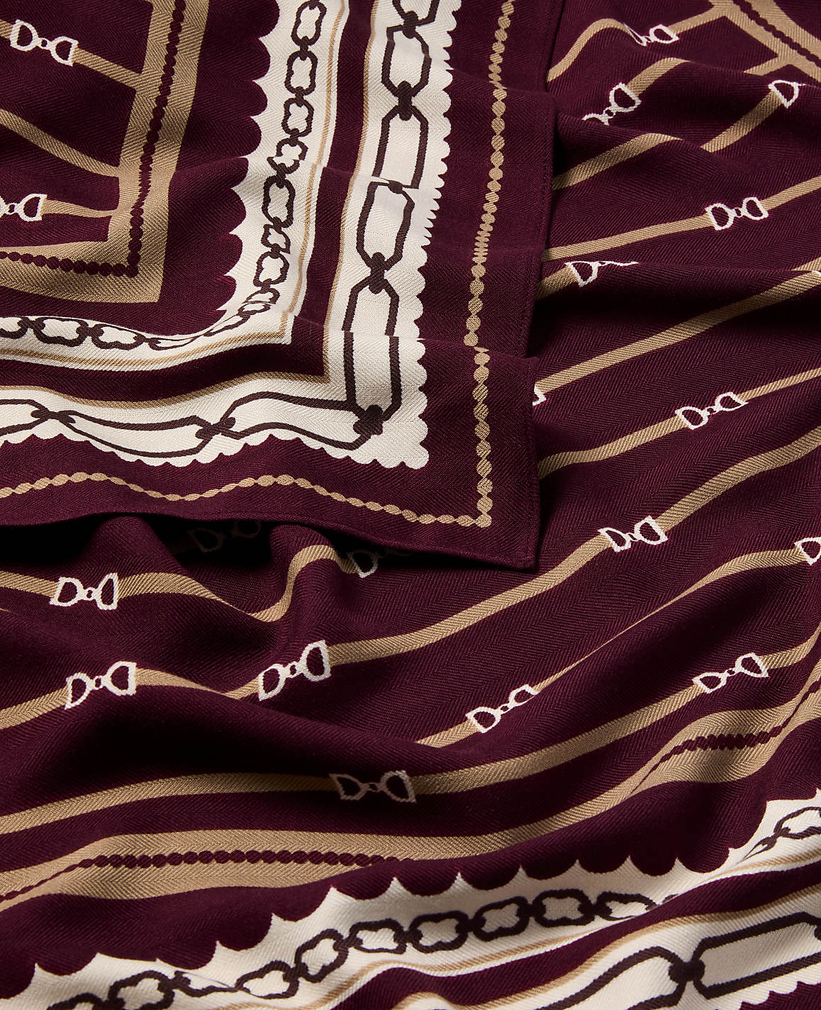 A folded burgundy scarf with a repeating equestrian bit pattern in beige and white is laid out, with a decorative border of chains and scallops visible on the left.