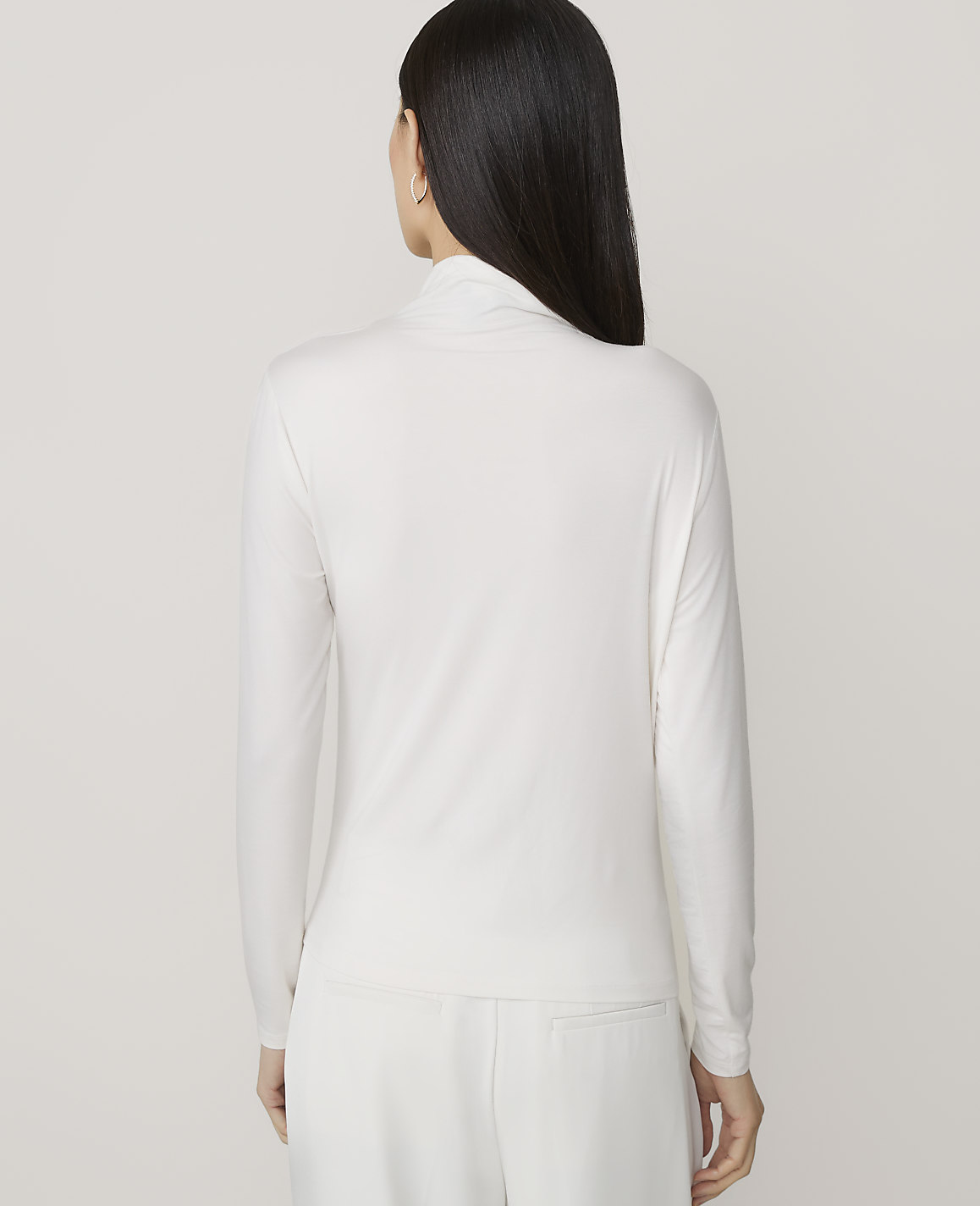 A woman in a white turtleneck and pants stands against a plain white background.
