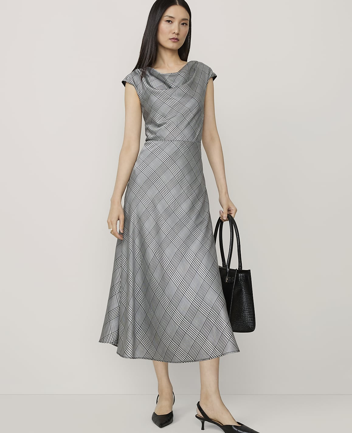 A woman in a grey plaid dress and black heels stands with a black tote bag against a plain, light grey background.
