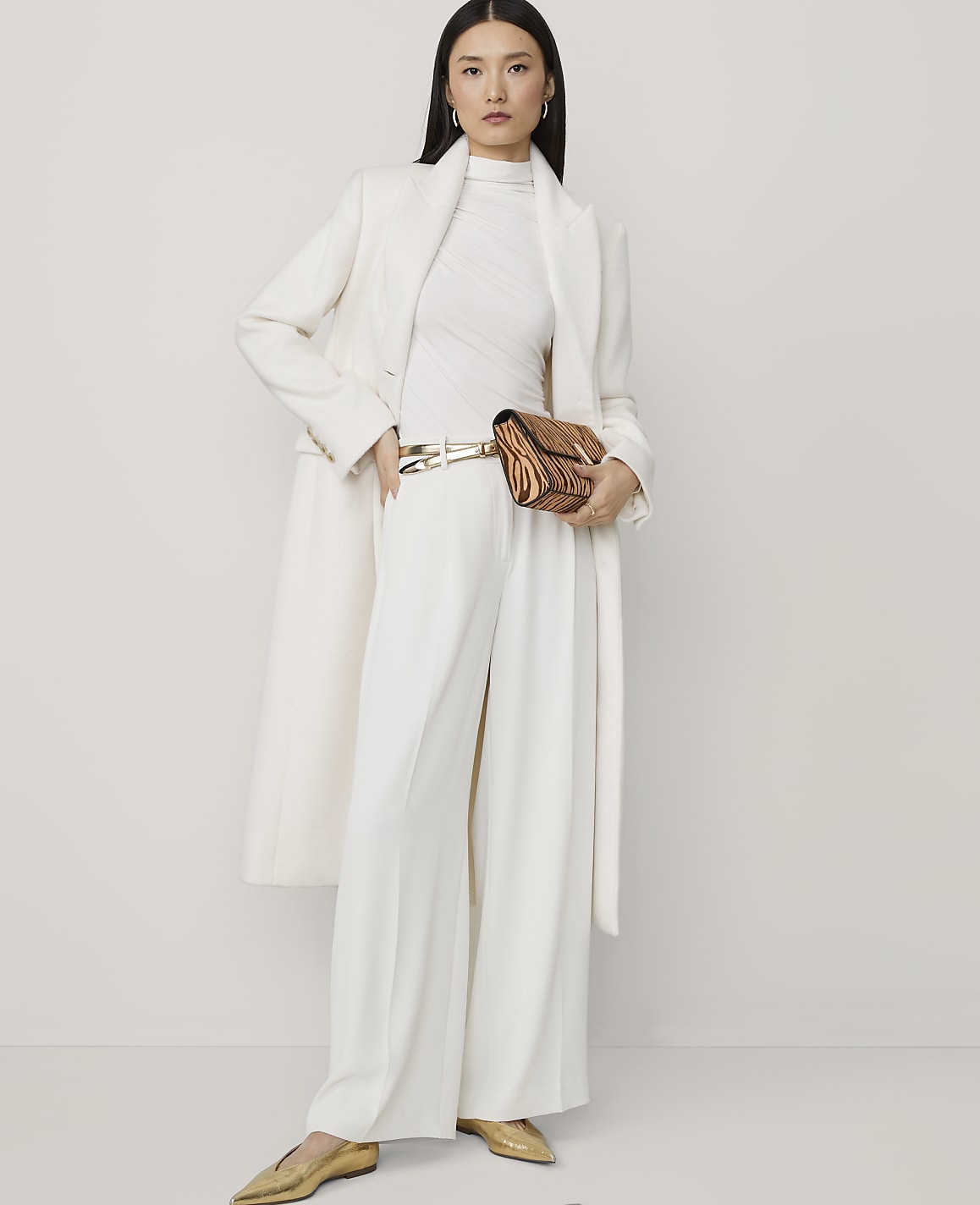 A woman in a white coat and pants poses against a plain white background, holding a patterned clutch.