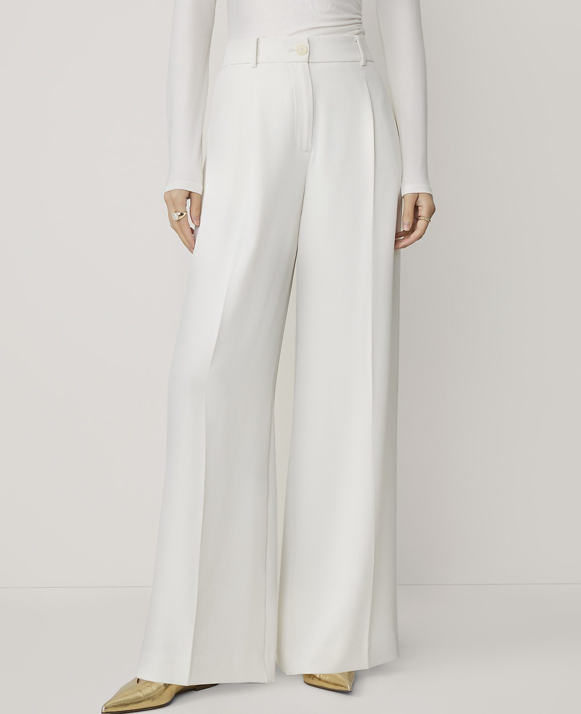 A person wearing white wide-leg pants and gold pointed-toe shoes stands against a plain white background.