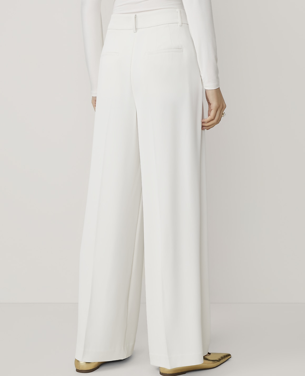 A person wearing white wide-leg pants and gold pointed-toe flats stands against a plain white background.