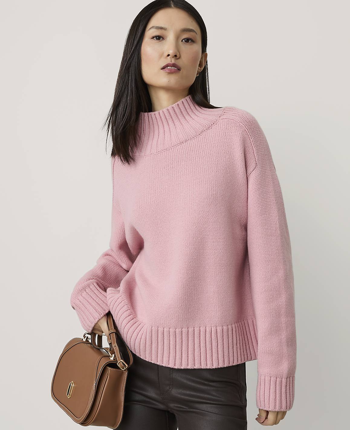 A woman in a pink sweater and dark pants holds a brown purse in front of a plain background.