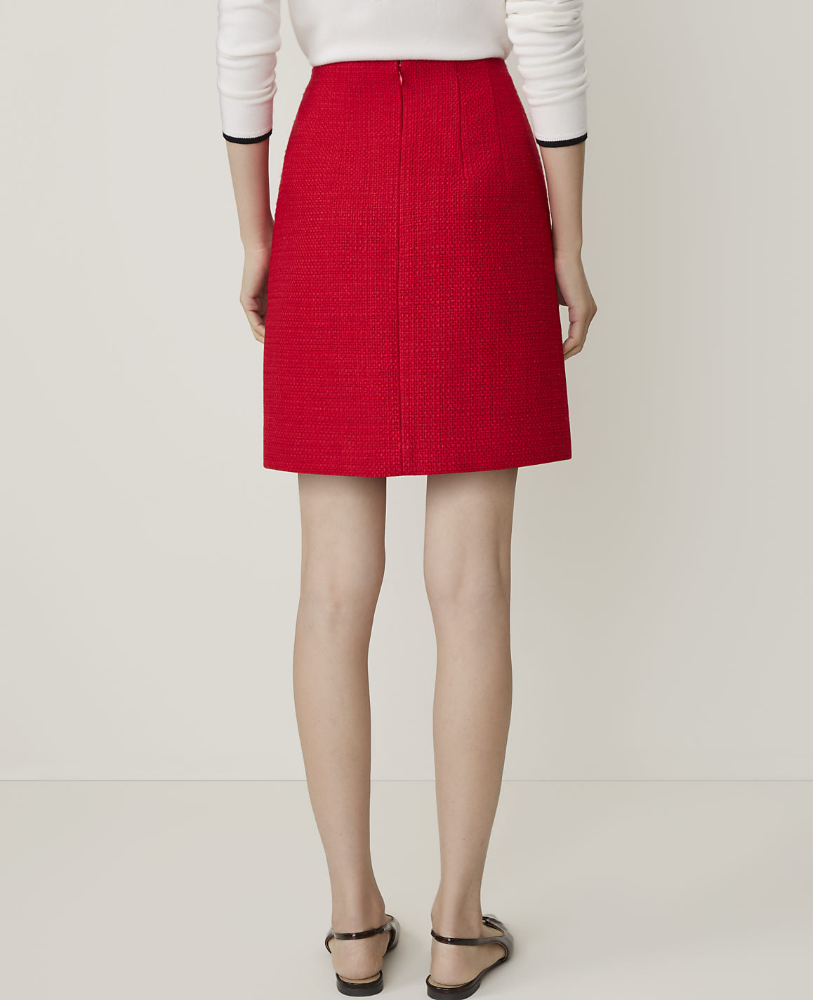 A woman in a red tweed skirt and black heels walks away from the camera against a plain white background.