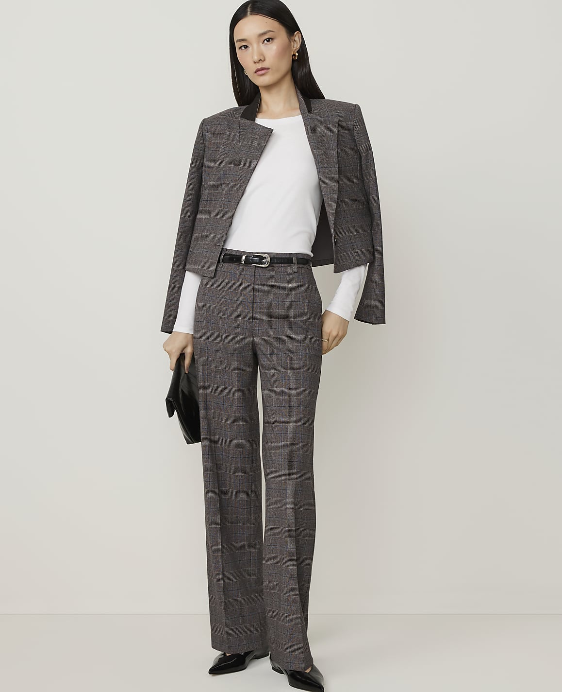 A woman in a grey plaid suit and black heels stands holding a dark woven clutch against a plain white background.