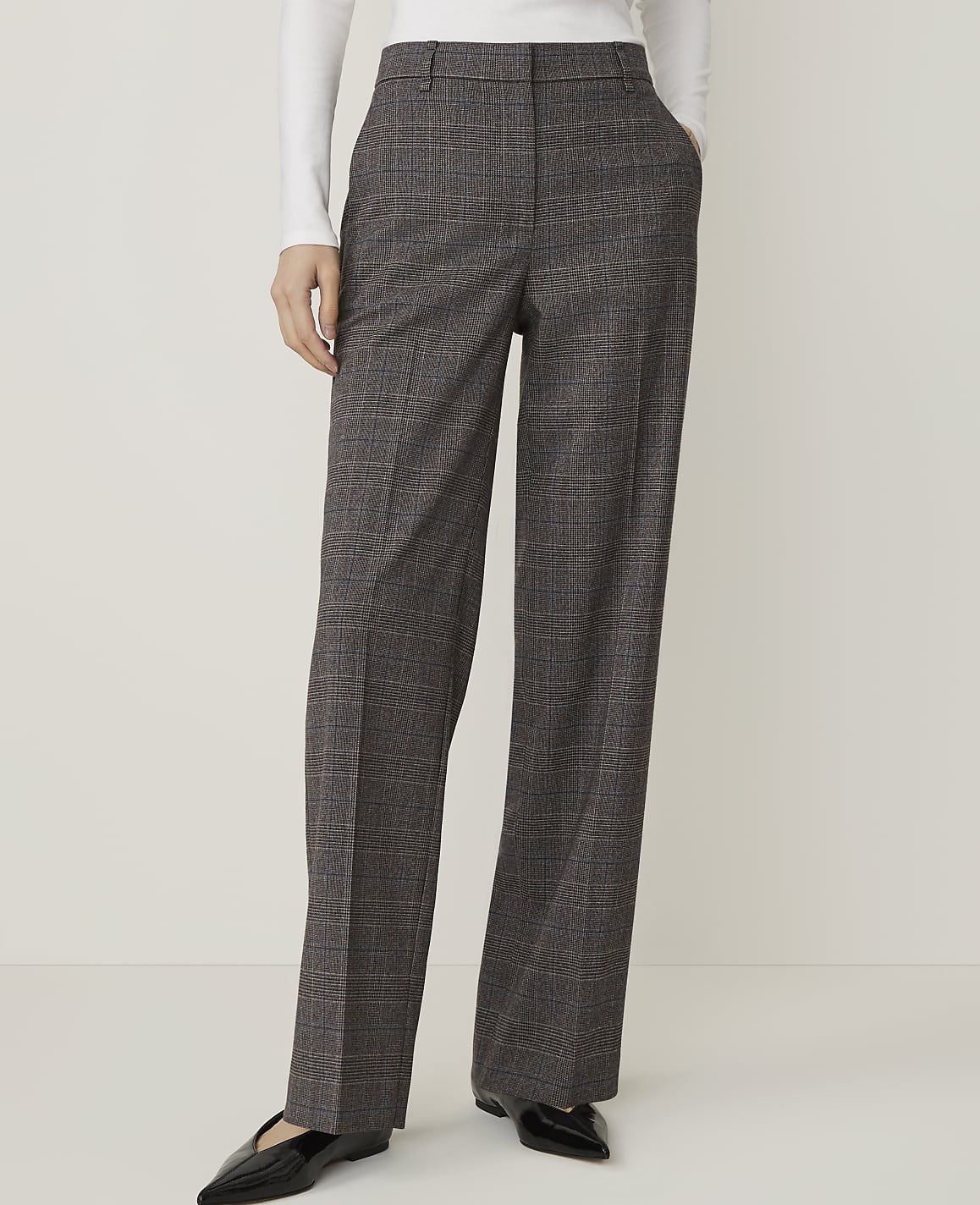 A person stands with hands in the pockets of their grey plaid wide-leg trousers against a plain white background.