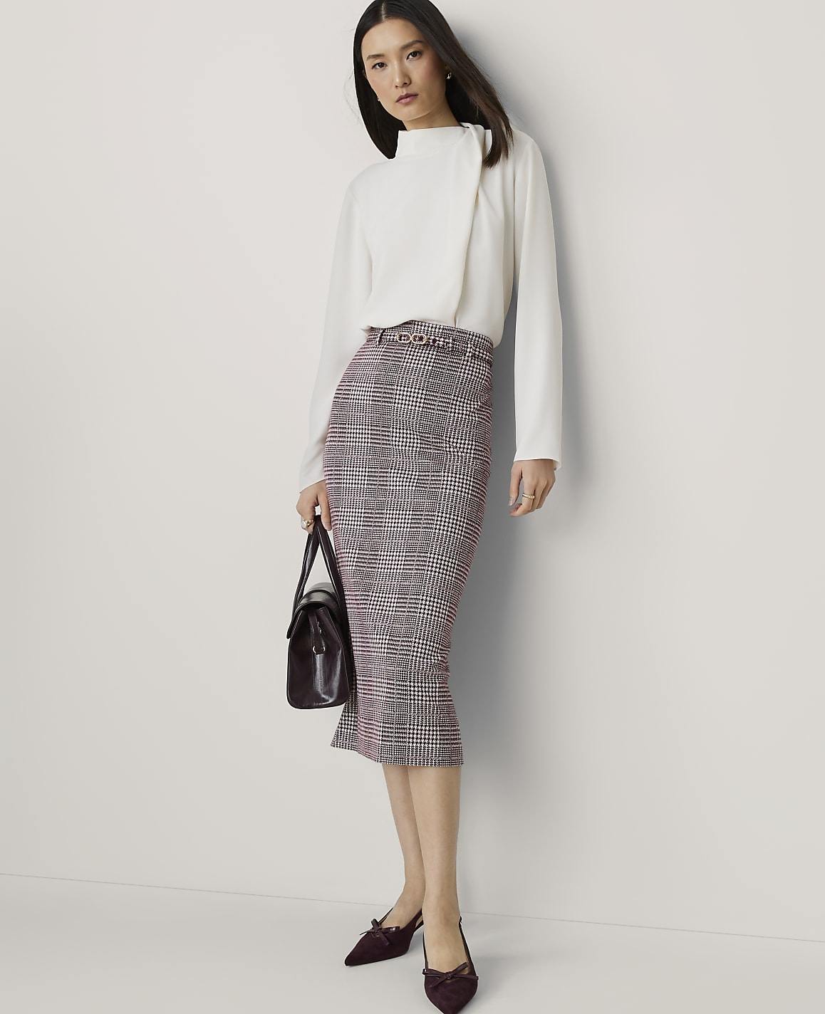 A woman in a white blouse and plaid skirt stands against a plain white background, holding a dark handbag.