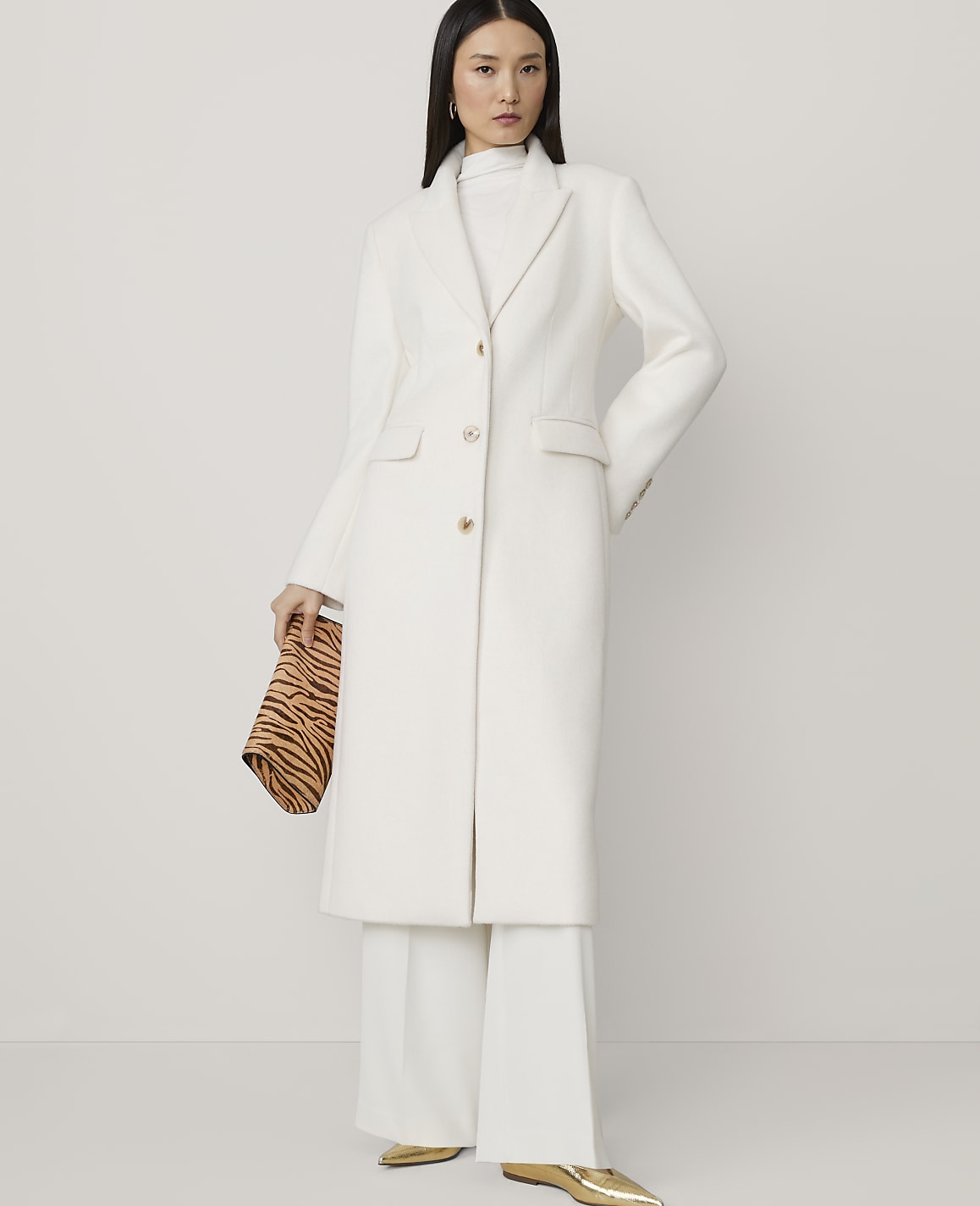 A woman in a white coat and wide-leg pants holds a tiger-print clutch against a plain white background.