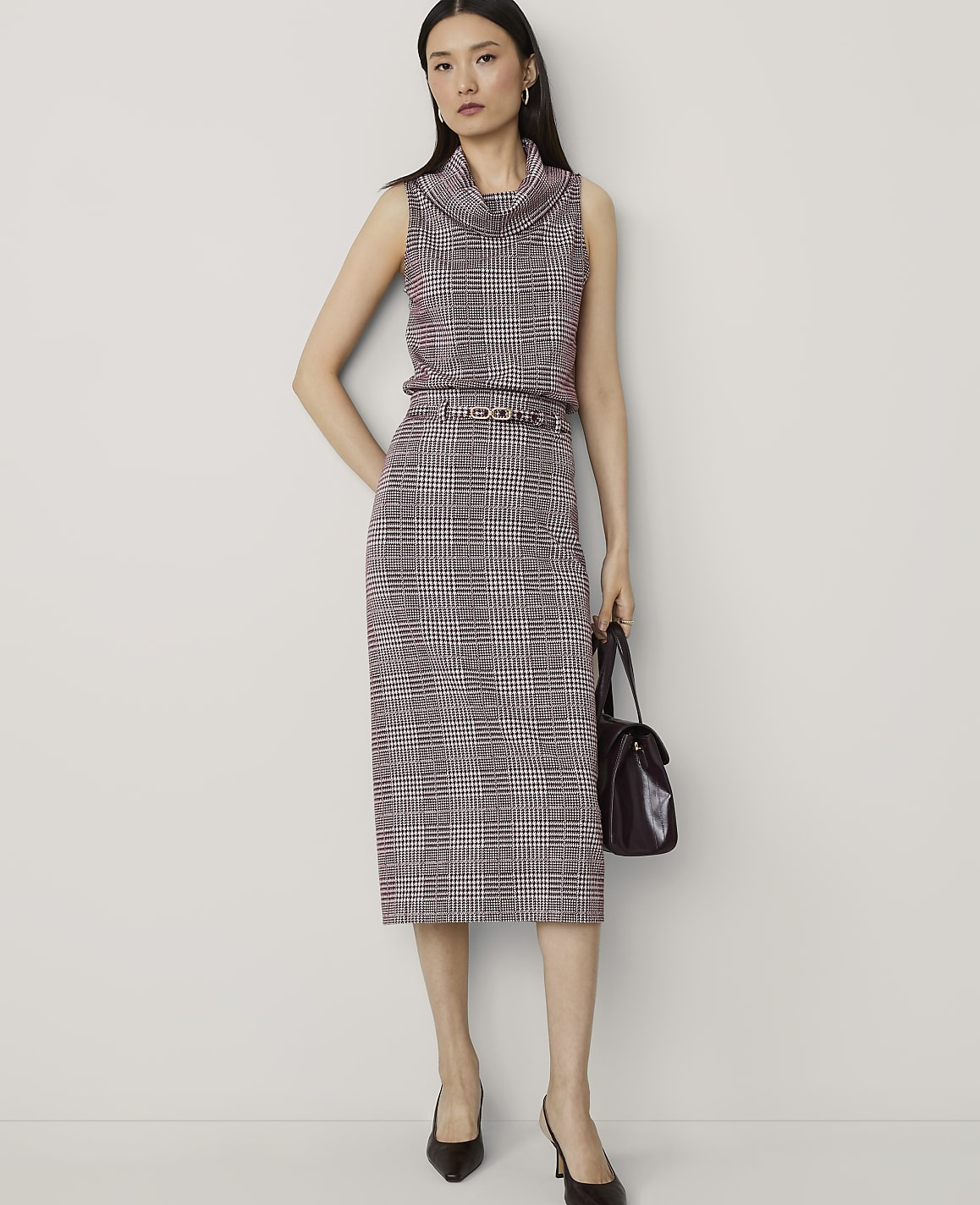 A woman in a plaid sleeveless turtleneck and skirt stands against a plain background, holding a dark handbag.