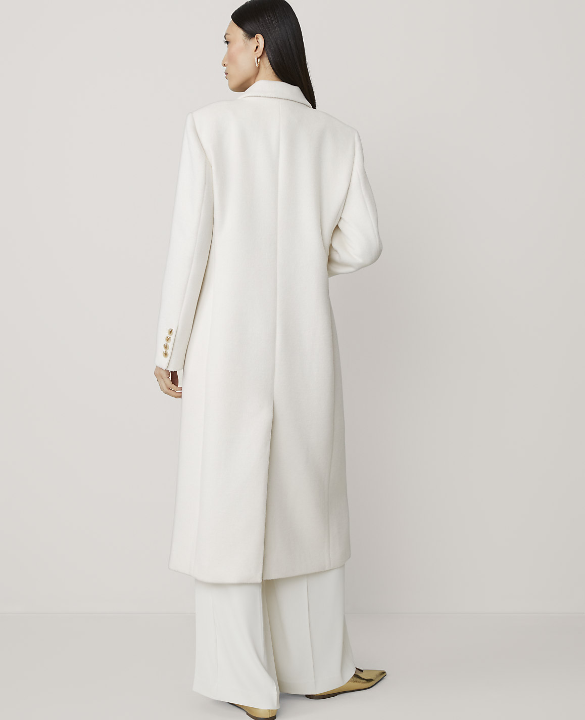 A woman in a long white coat and wide-leg pants stands against a plain, light background.
