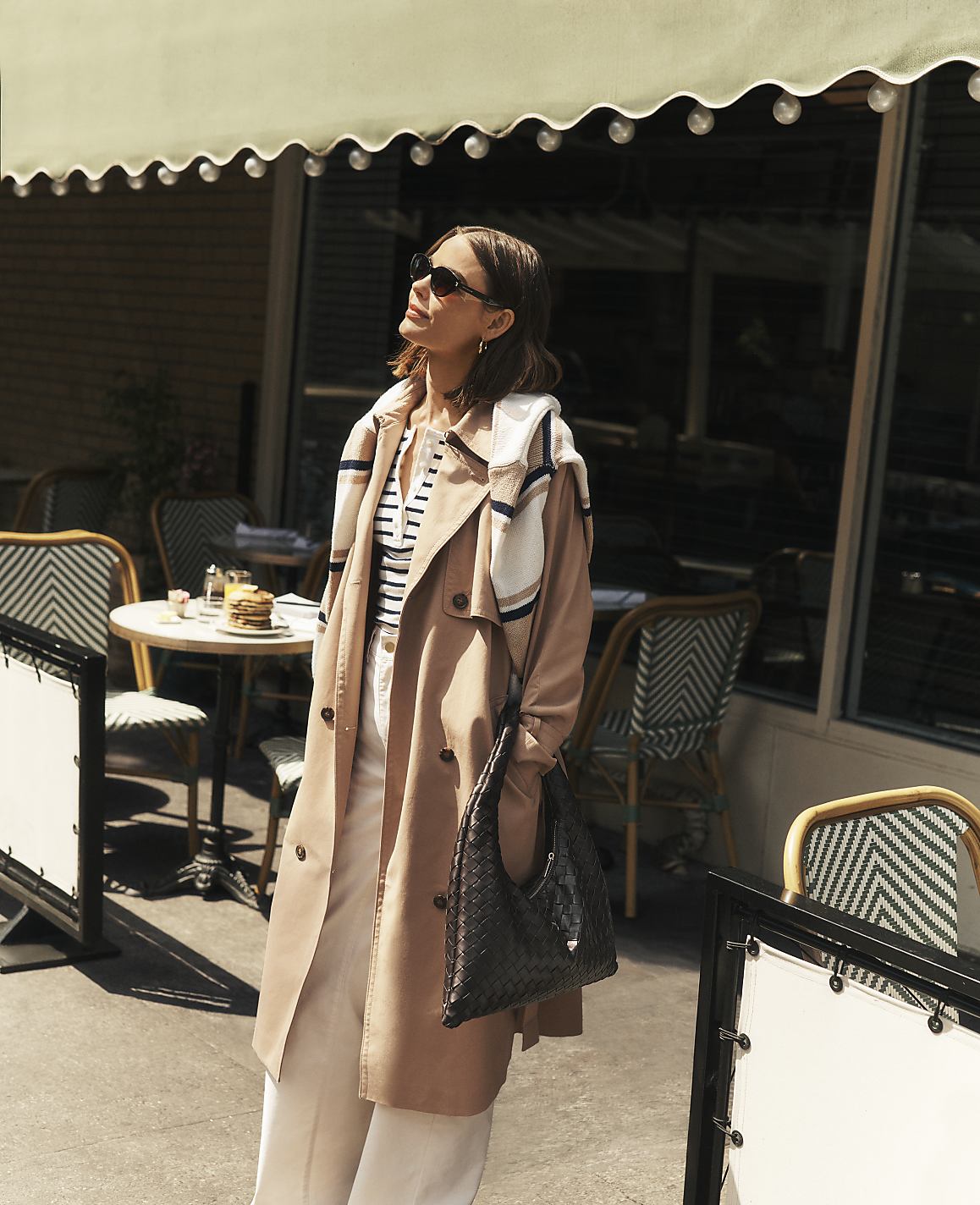 A woman in a trench coat stands in front of a cafe with tables and chairs visible in the background.