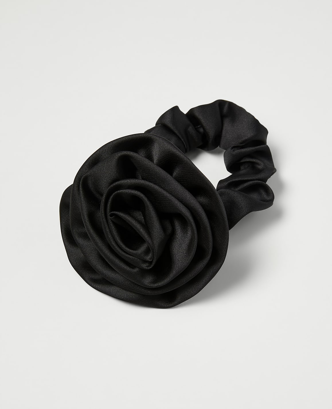 A black satin scrunchie with a rose embellishment rests on a plain white background.