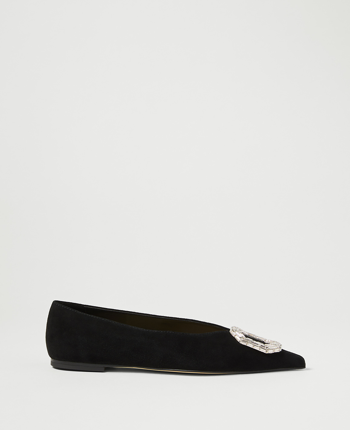 A black suede flat shoe with a jeweled buckle sits on a plain white background.