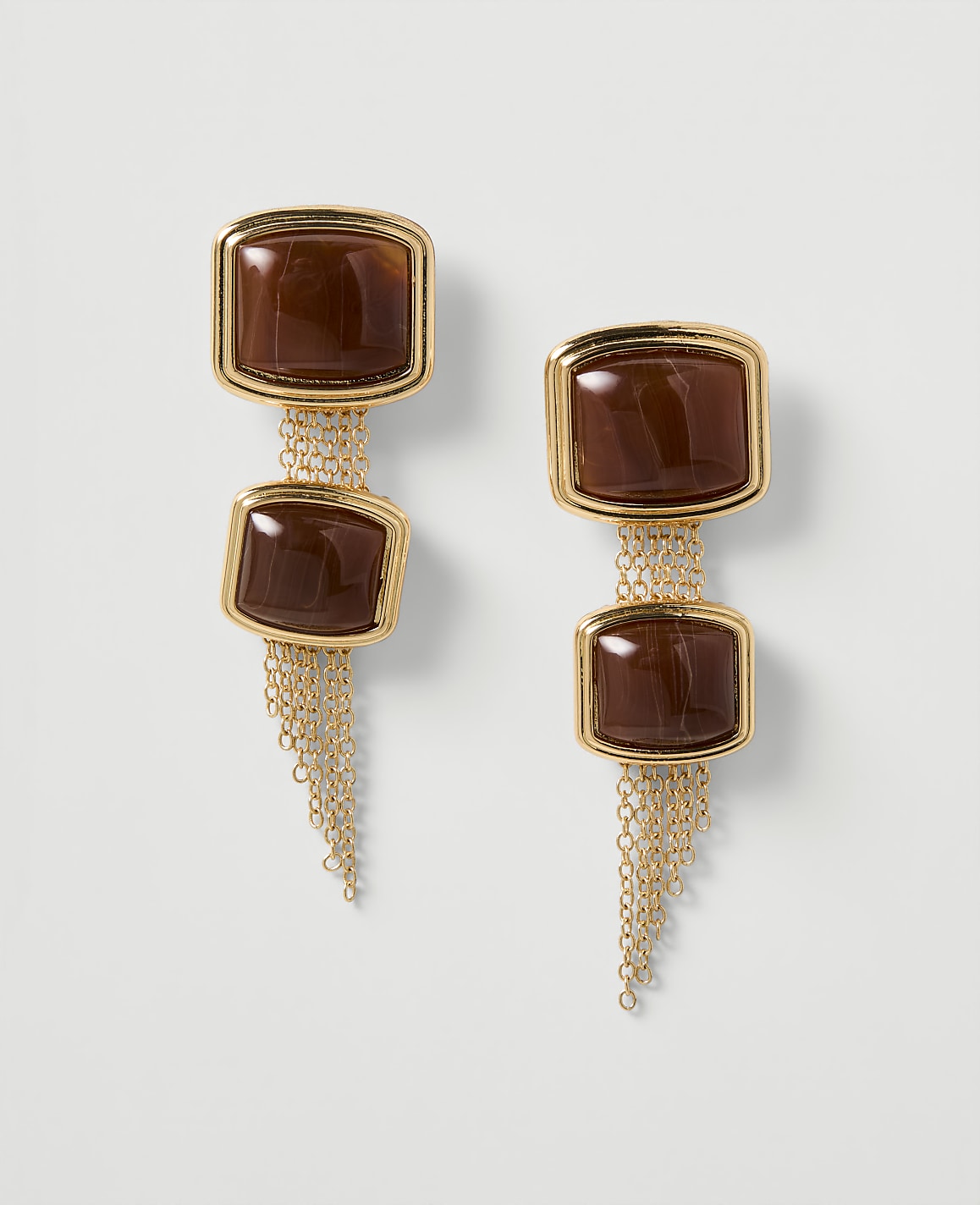 A pair of gold earrings with brown square stones and delicate chain tassels hang against a plain white background.