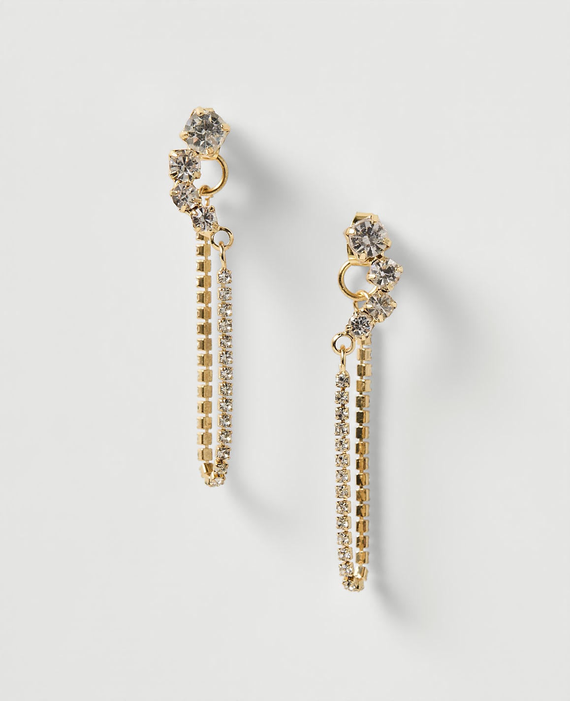 A pair of gold dangle earrings with sparkling rhinestones and chain fringe hang against a plain white background.
