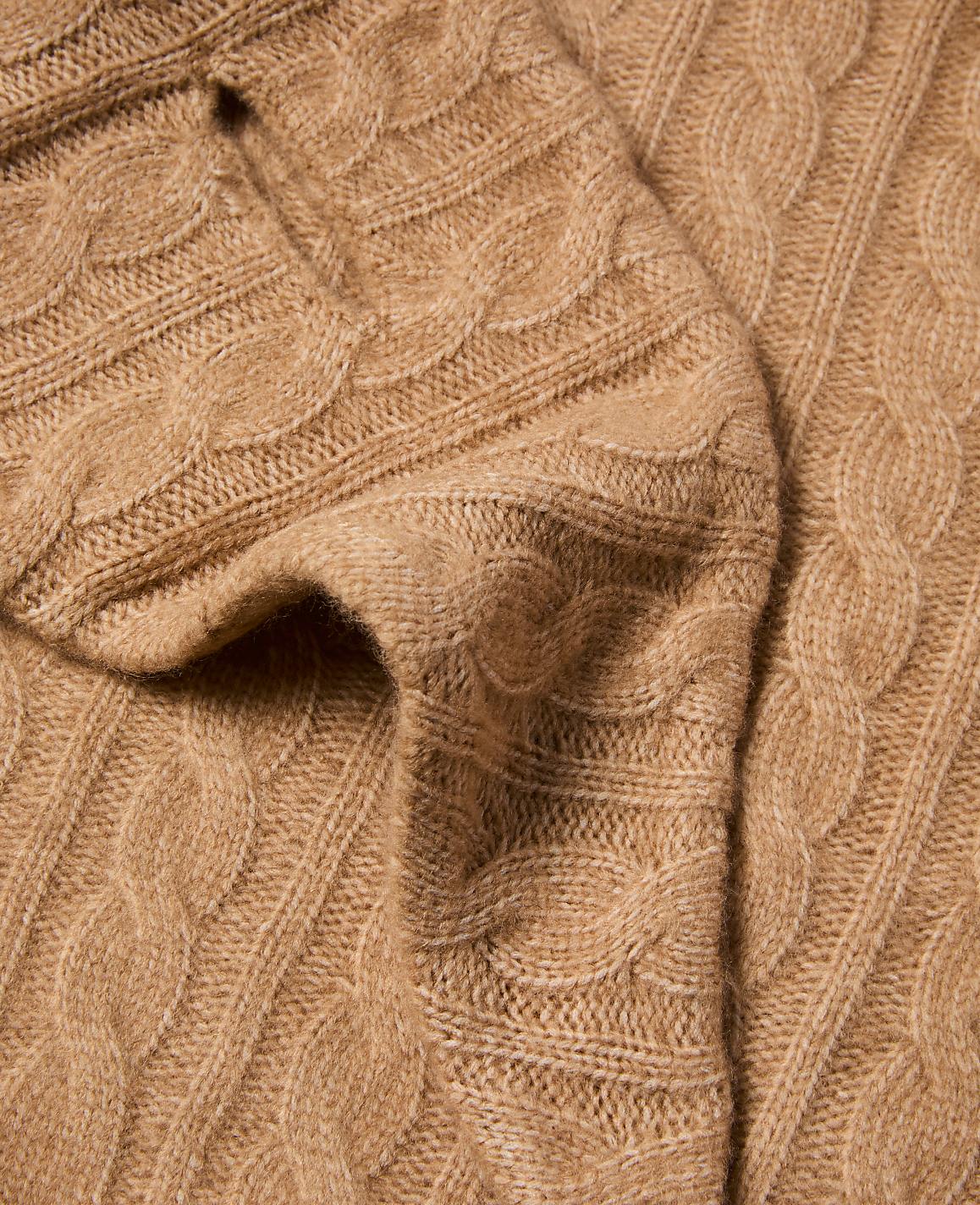 A close-up view of a soft, camel-colored cable-knit sweater fills the frame, with its textured weave and gentle folds dominating the foreground and background.