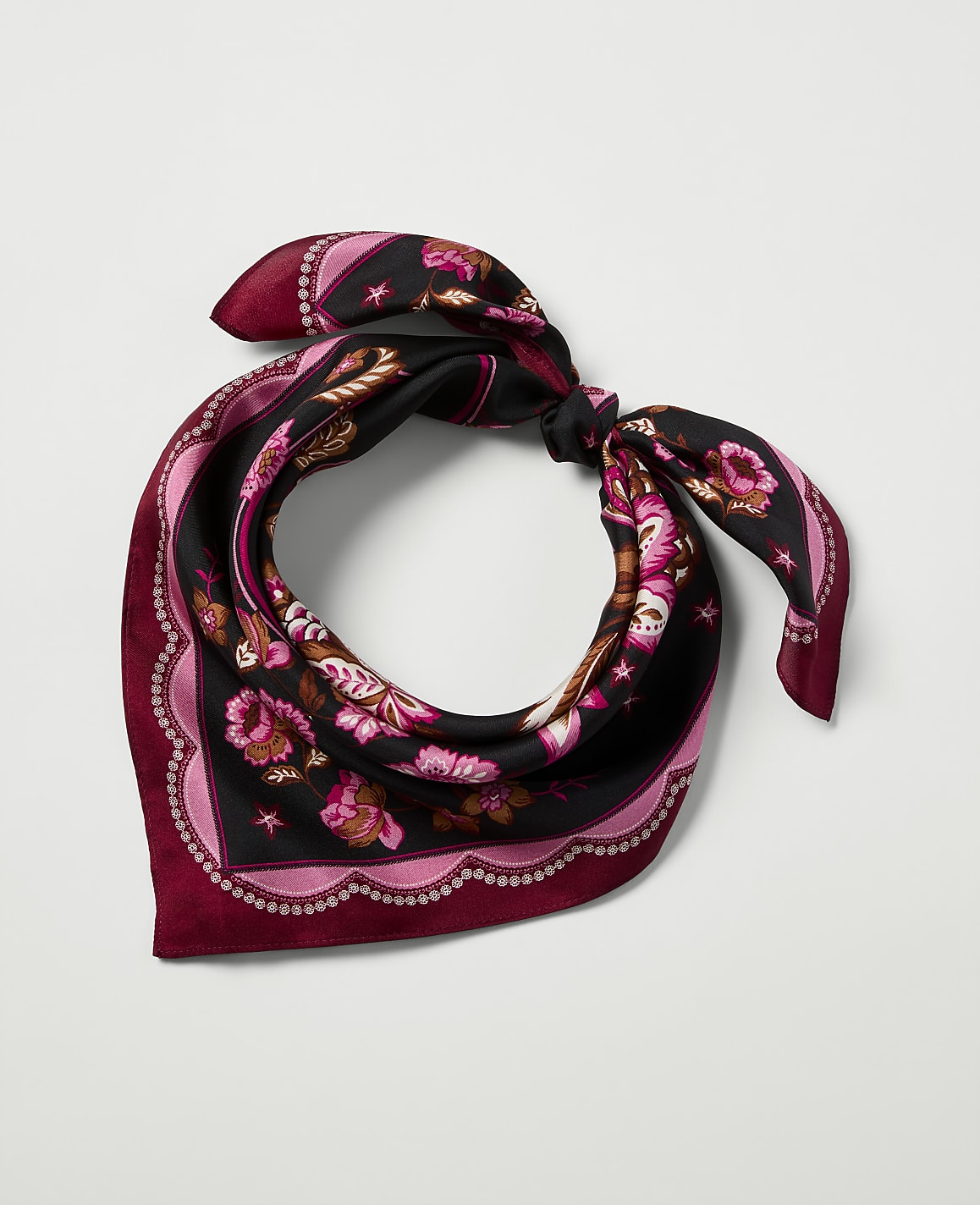 A black silk scarf with a floral pattern and pink scalloped border is tied in a knot against a plain white background.