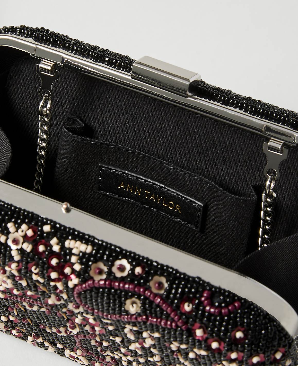 A black beaded clutch with a floral design in the foreground is open to reveal a black interior with an Ann Taylor label in the background.