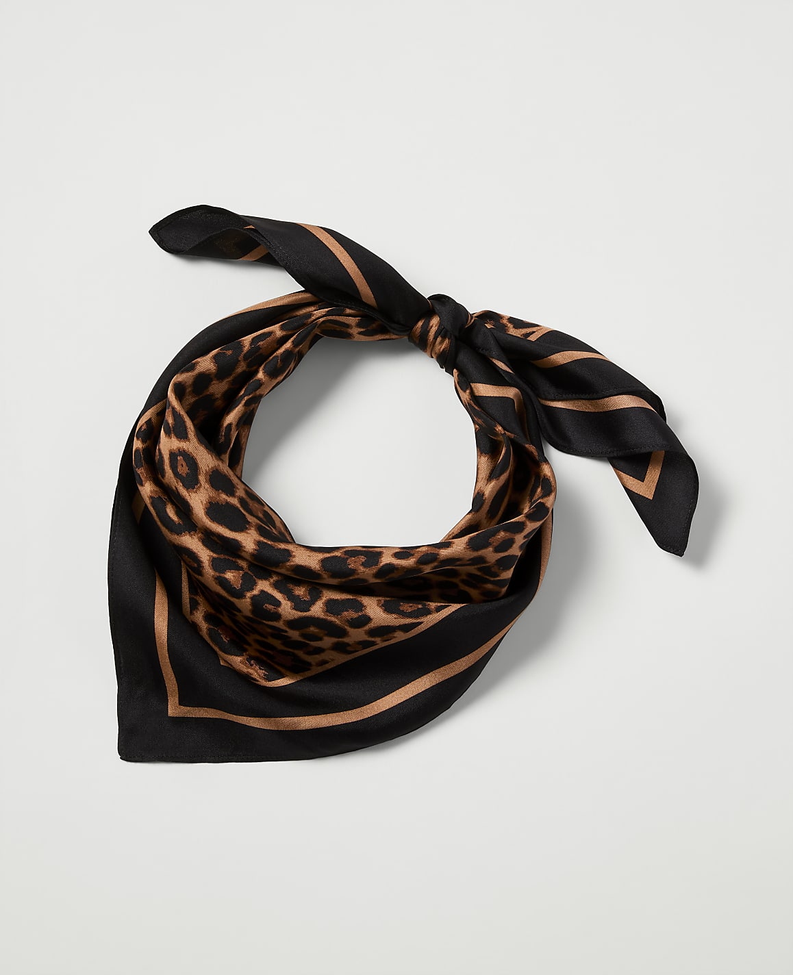 A leopard print silk scarf with black and tan borders is tied in a knot against a plain white background.