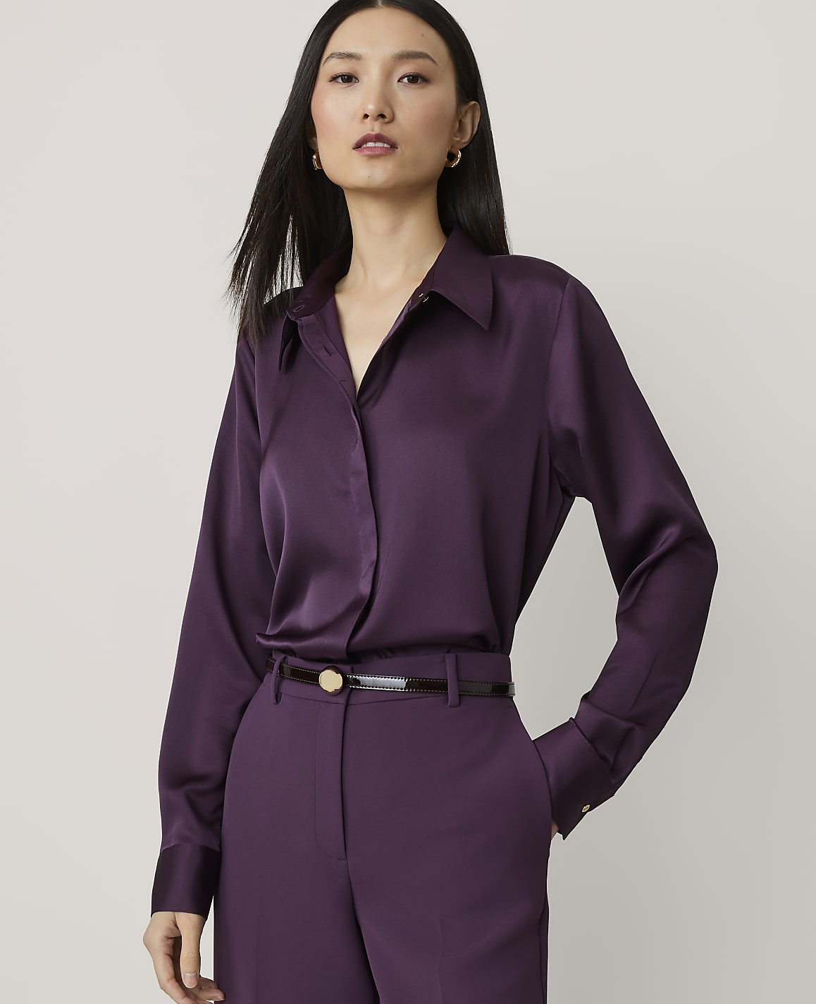 A woman in a purple satin blouse and matching pants stands against a plain, light gray background.