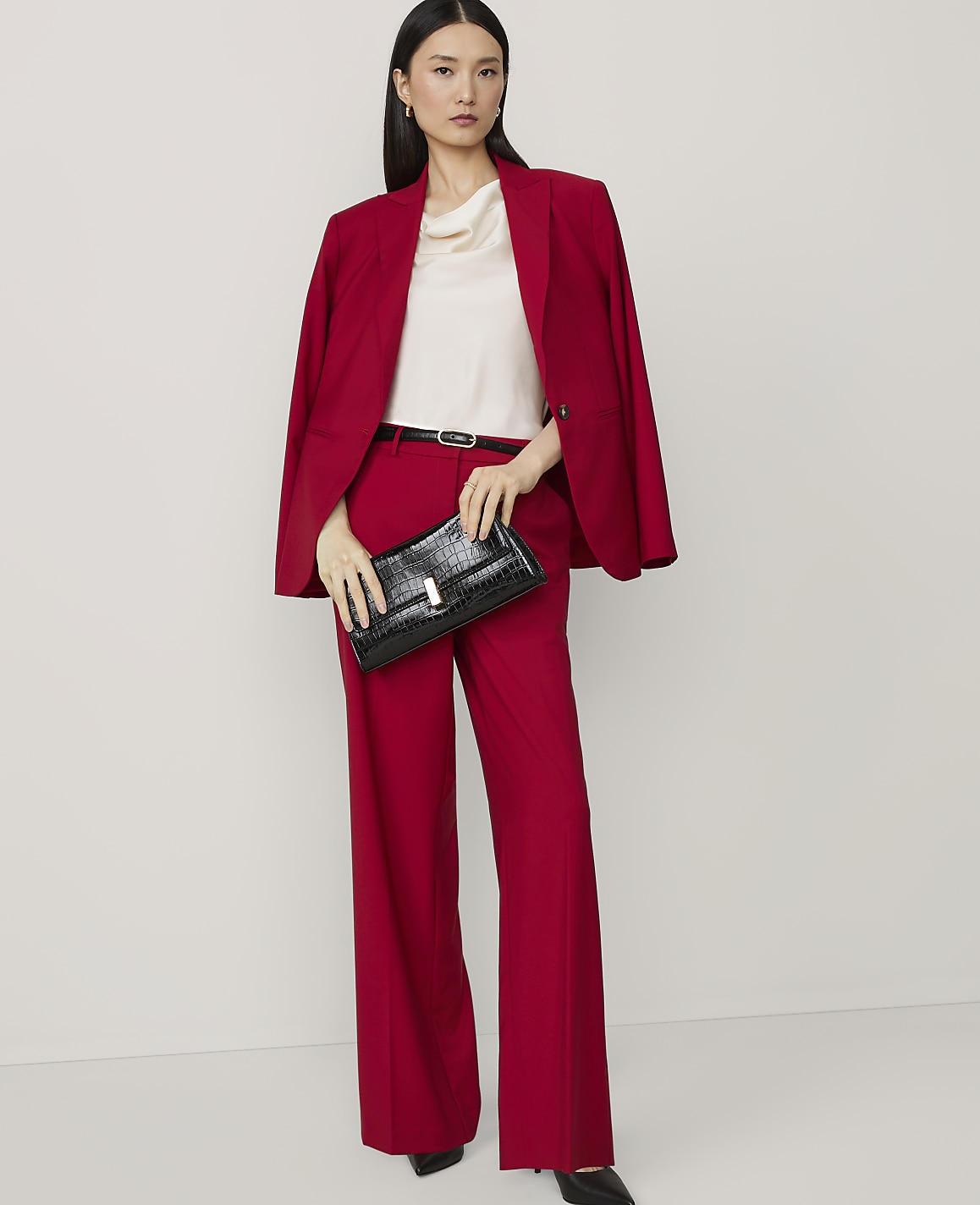 A woman in a red suit and cream top holds a black clutch in front of a plain white background.