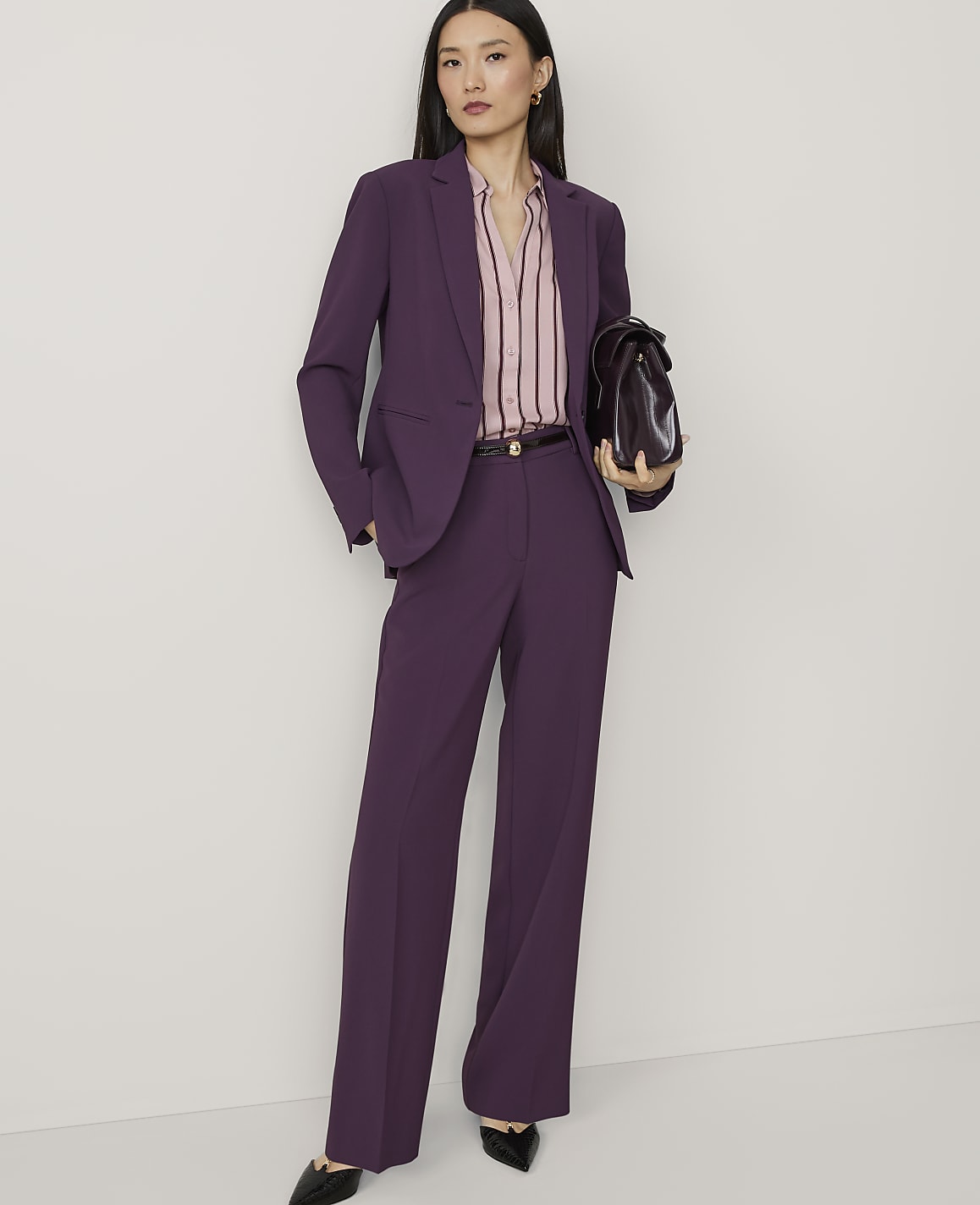 A woman in a purple pantsuit stands against a plain white background, holding a dark brown briefcase.