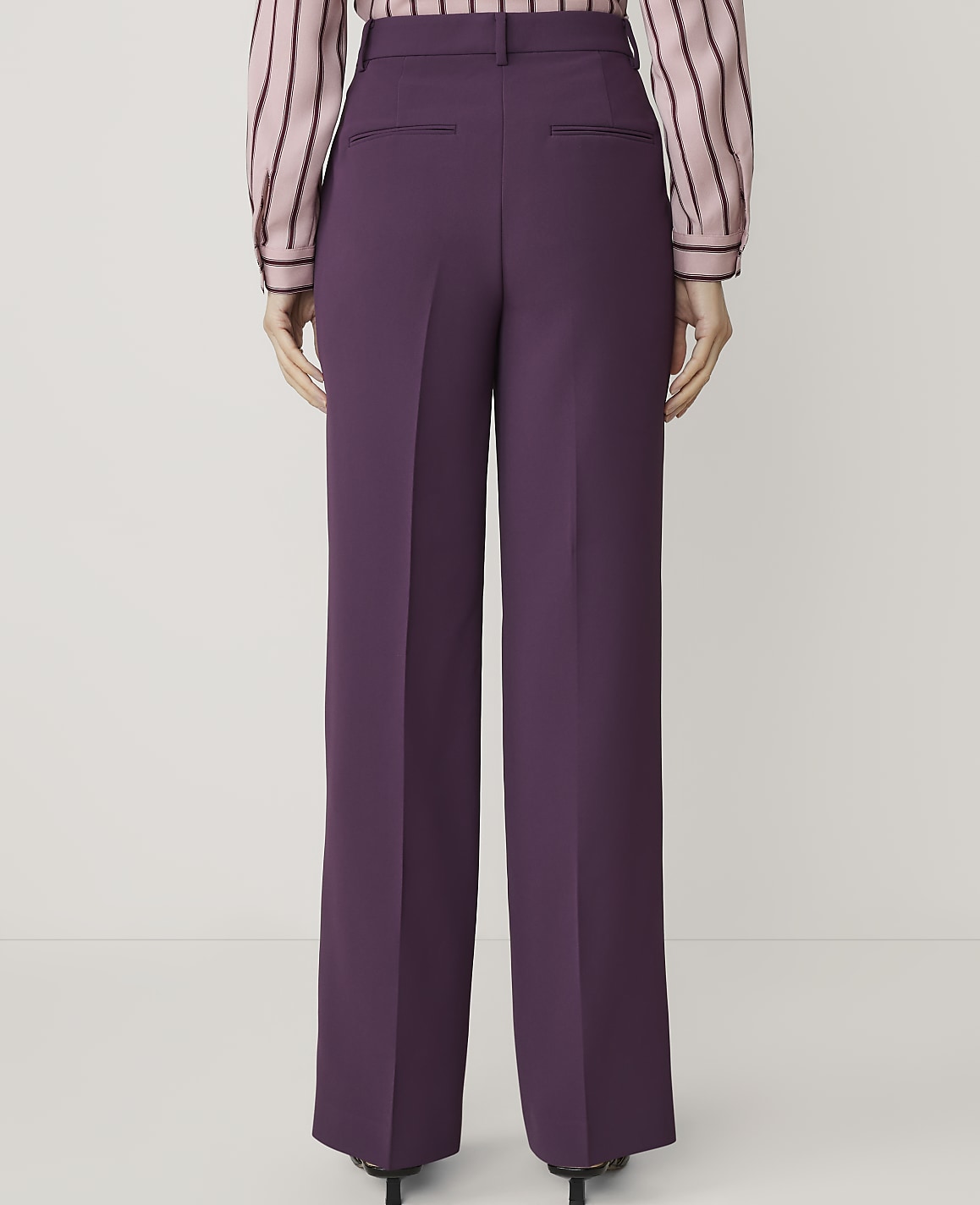 A person wearing purple wide-leg trousers and a striped pink and black blouse stands against a plain white background.