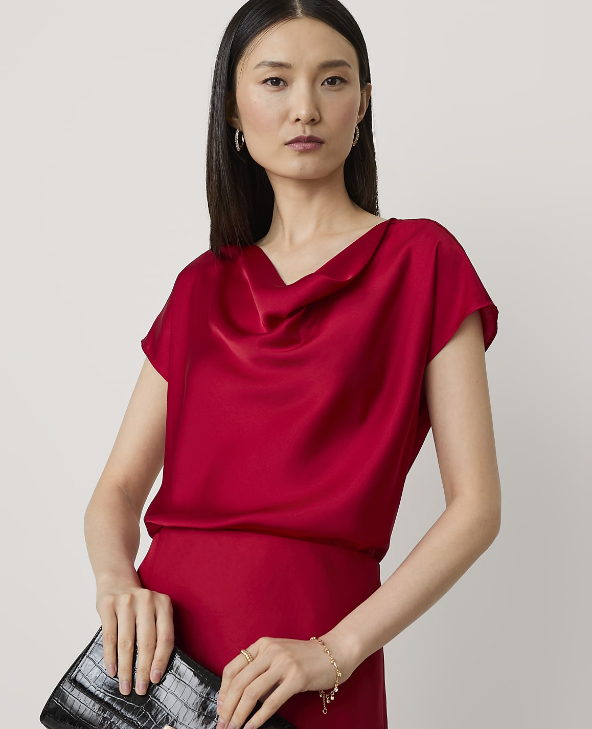 A woman in a red cowl-neck top and holding a black clutch stands against a plain white background.