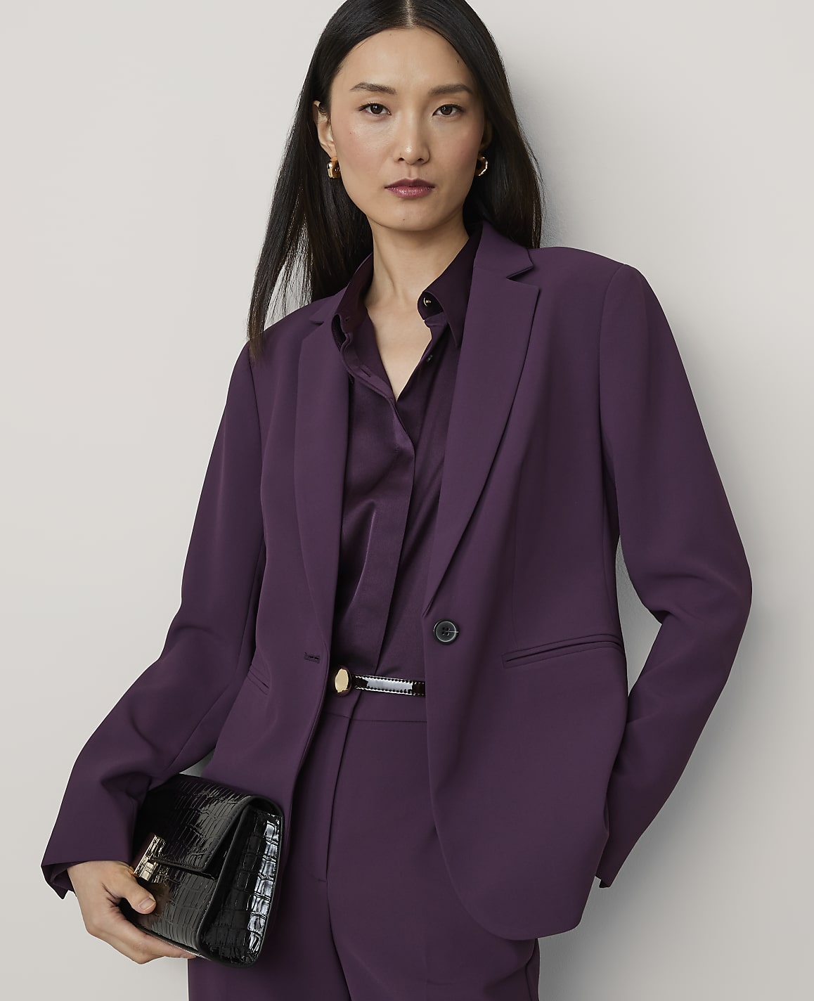 A woman in a purple suit holds a black clutch in front of a plain gray background.