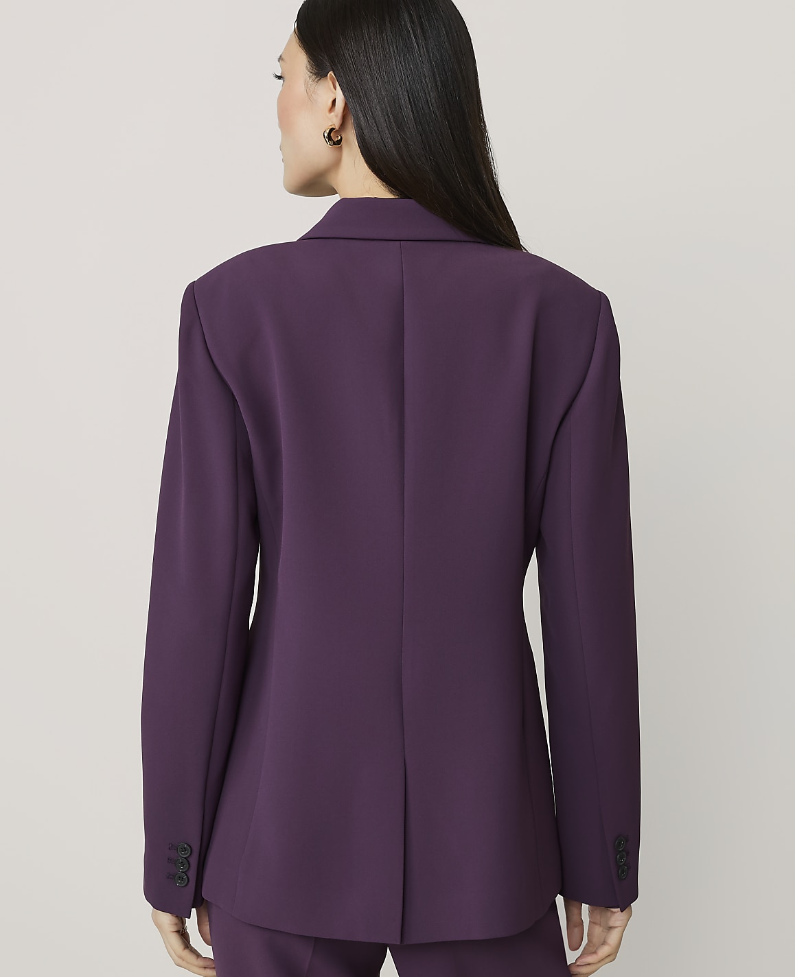 A woman in a purple blazer stands against a plain, light gray background.