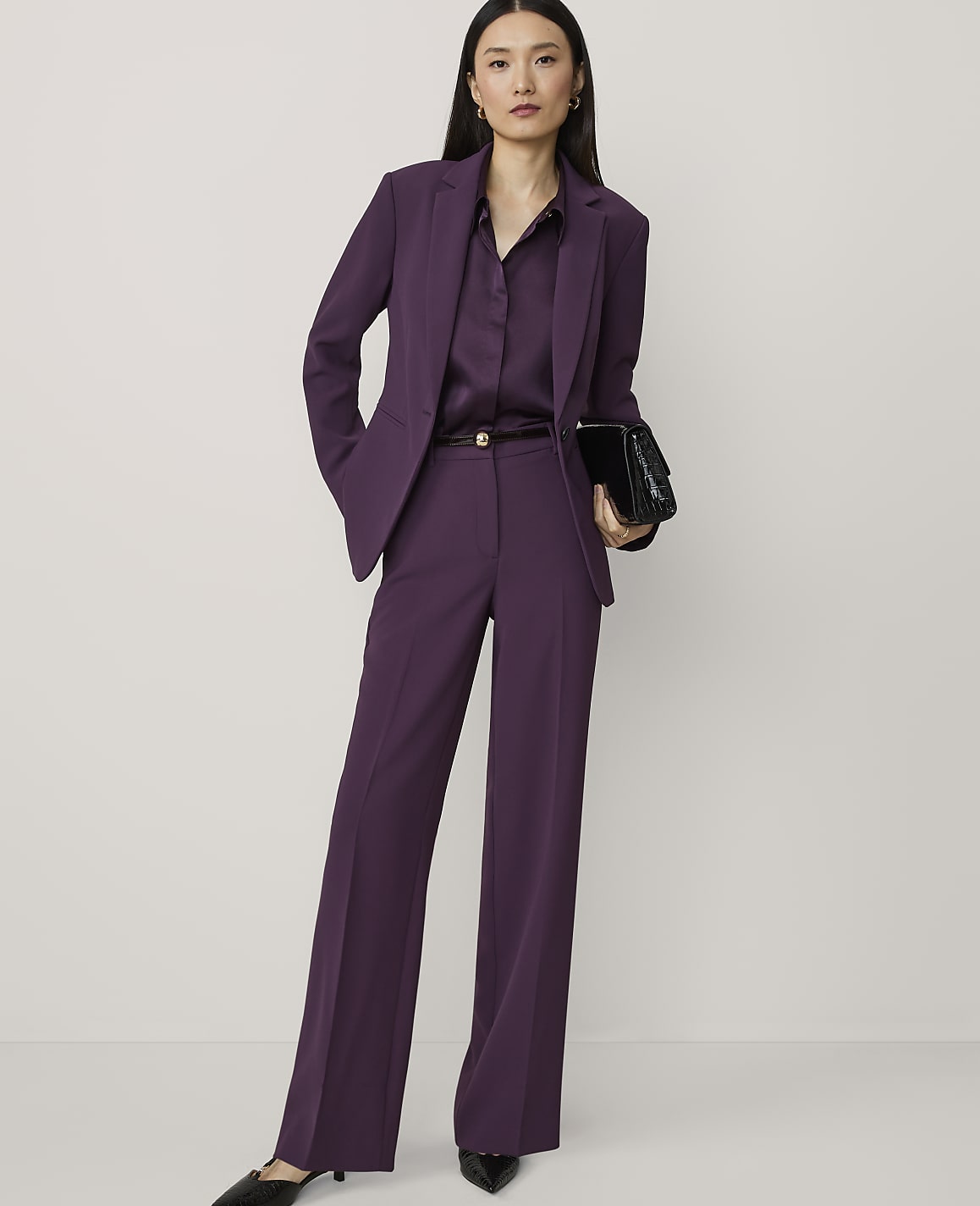 A woman in a purple pantsuit stands against a plain white background, holding a black clutch.