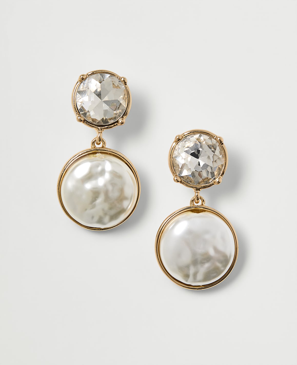 A pair of gold-toned drop earrings, each featuring a clear crystal stud above a lustrous pearl disc, are presented against a plain white background.