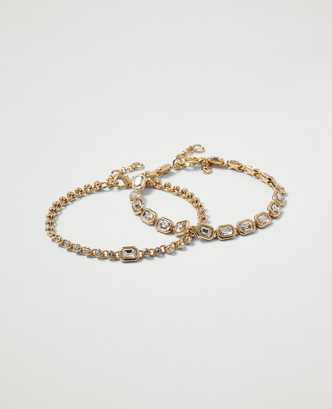 Two delicate gold bracelets, one with a simple chain and the other adorned with clear stones, rest against a plain white background.