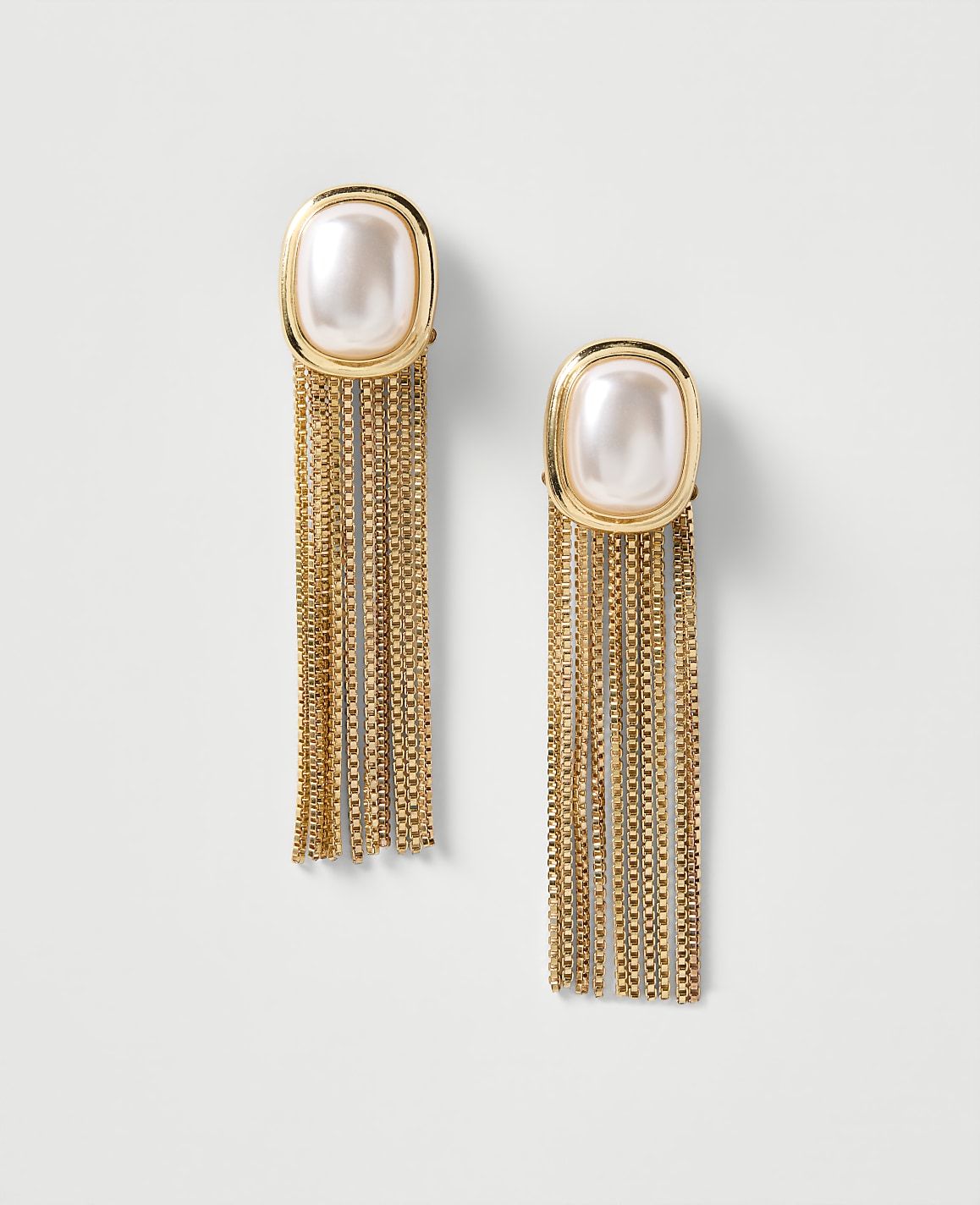 A pair of gold earrings with pearl tops and long, shimmering fringe hang against a plain, light background.